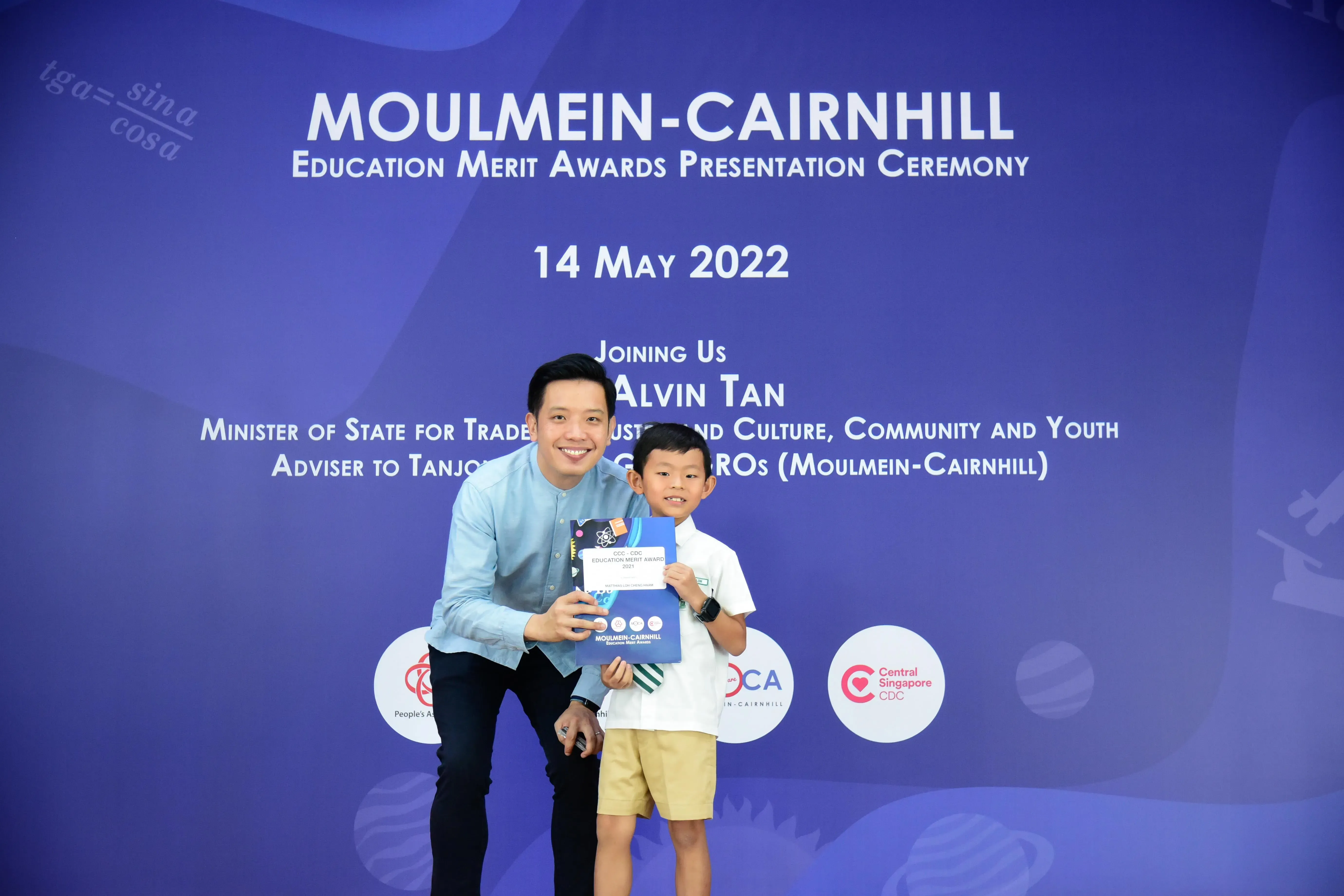 A man and a young boy hold an award certificate in front of a Moulmein-Cairnhill Education Merit Awards backdrop.