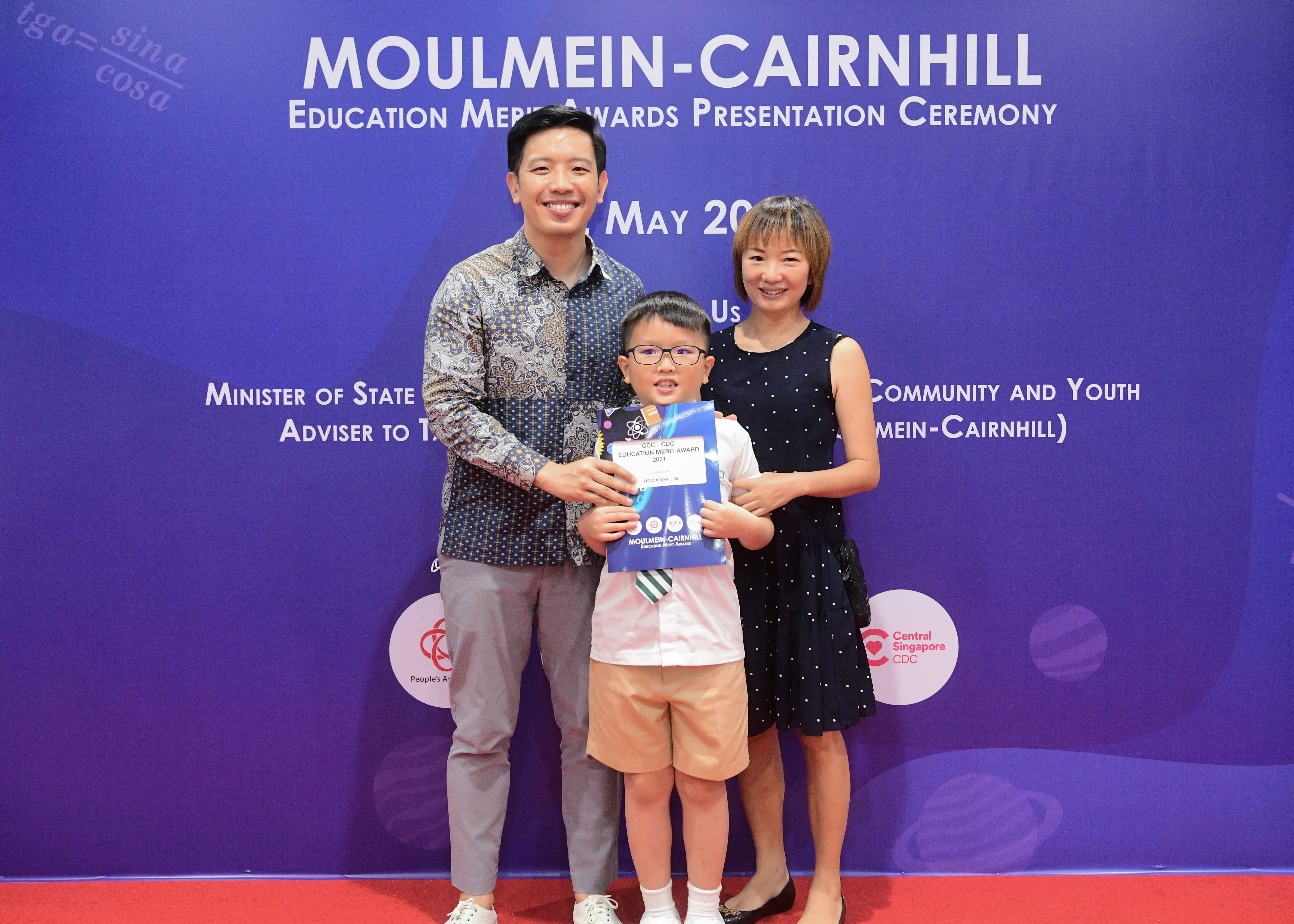 Two adults and a boy holding an award certificate pose before the Moulmein-Cairnhill Education Merit Awards banner.