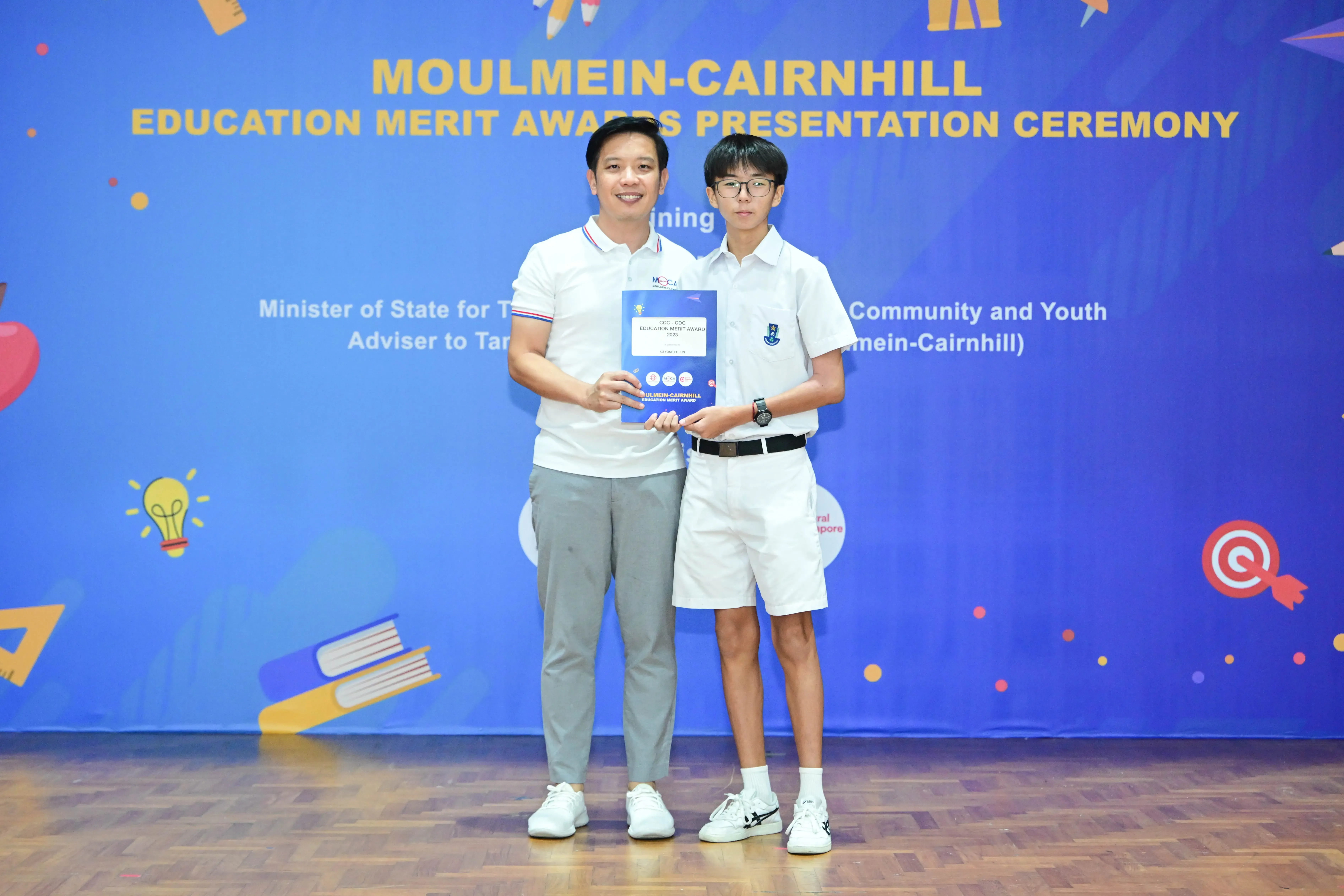Two people hold a certificate together in front of a Moulmein-Cairnhill Education Merit Awards banner.