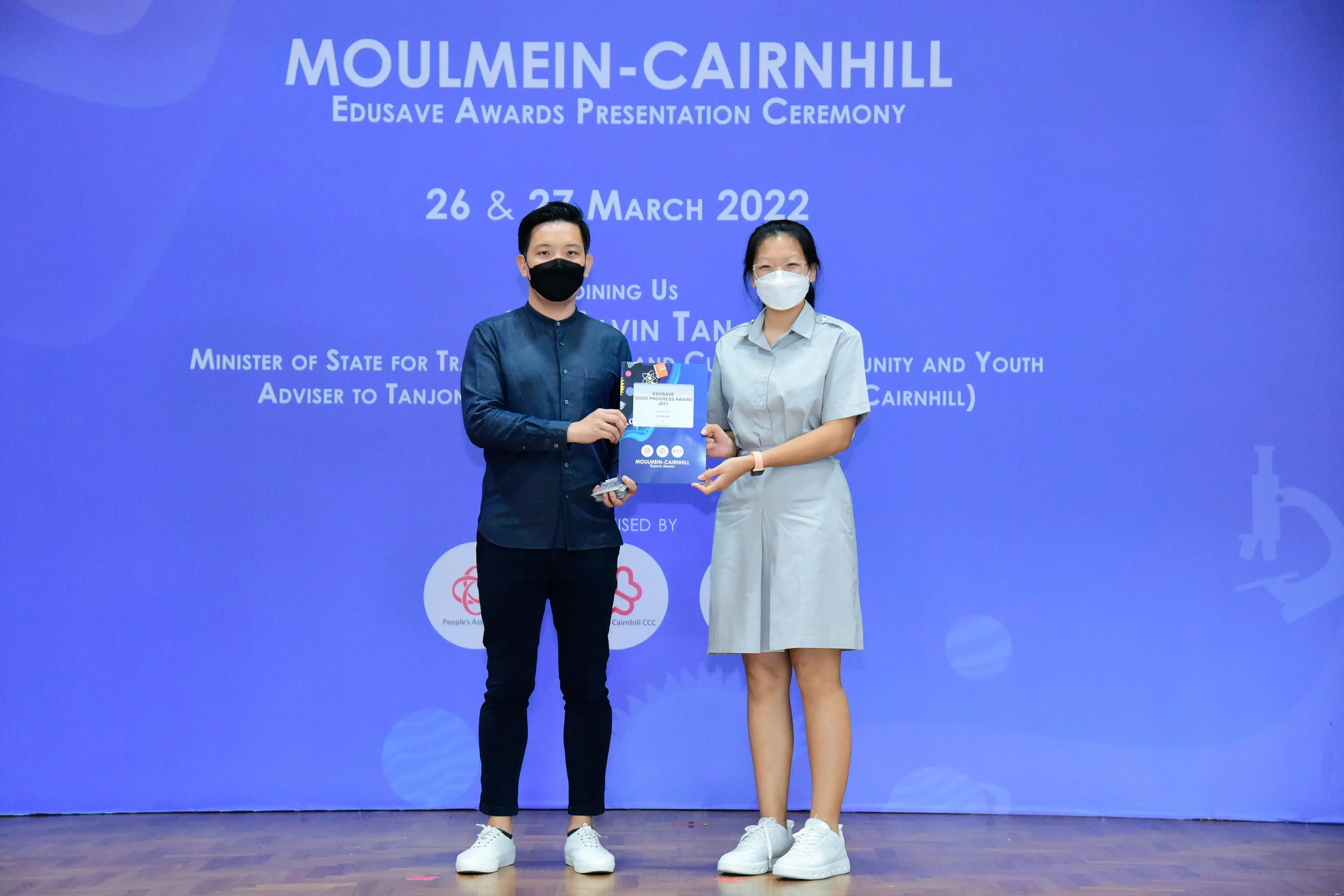 Two masked people hold an award at the Moulmein-Cairnhill Edusave Awards Presentation Ceremony, March 2022.