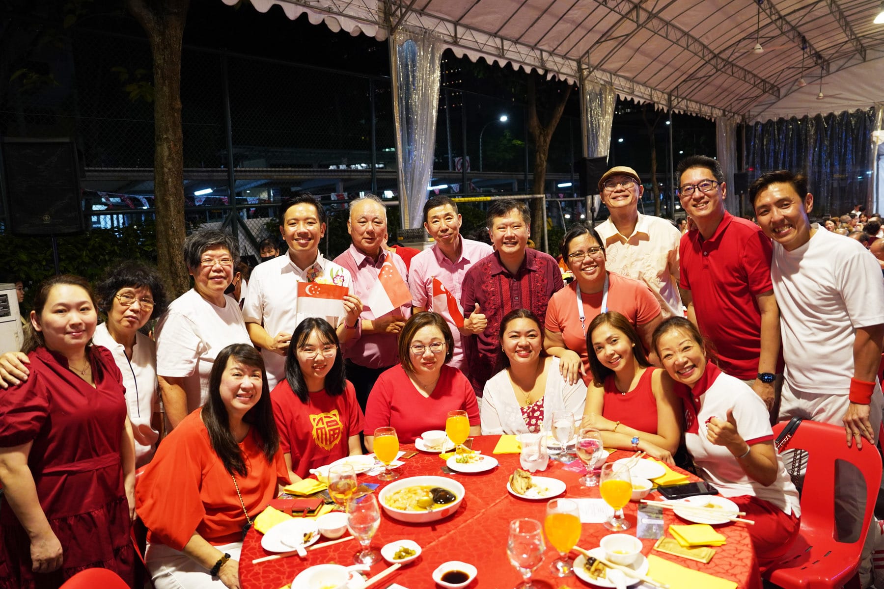 A large group of people in red and white outfits pose together at an outdoor dinner event, with Singapore flags visible.