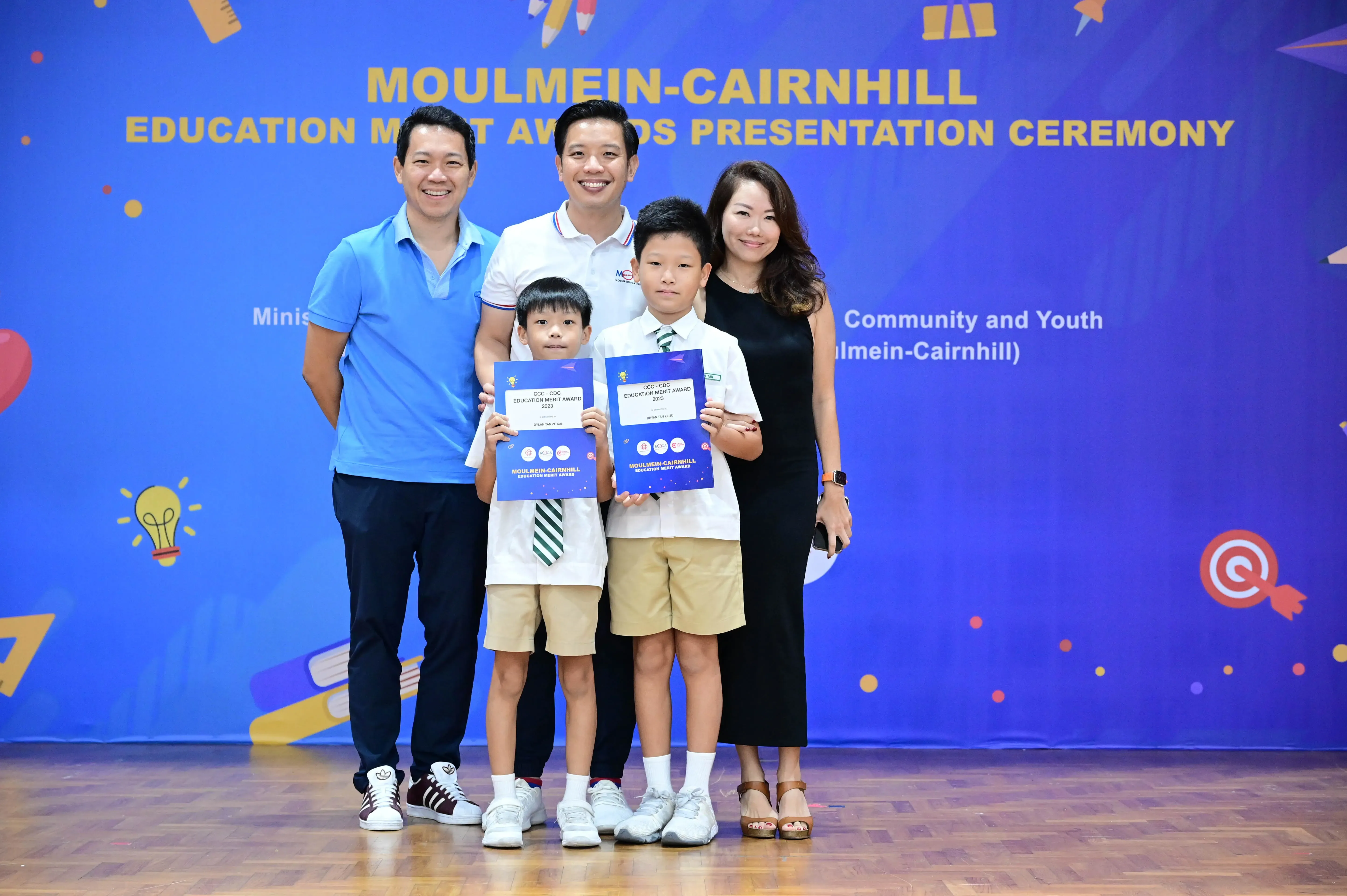 Two boys holding merit award certificates pose with three adults at the Moulmein-Cairnhill Education Merit Awards ceremony.