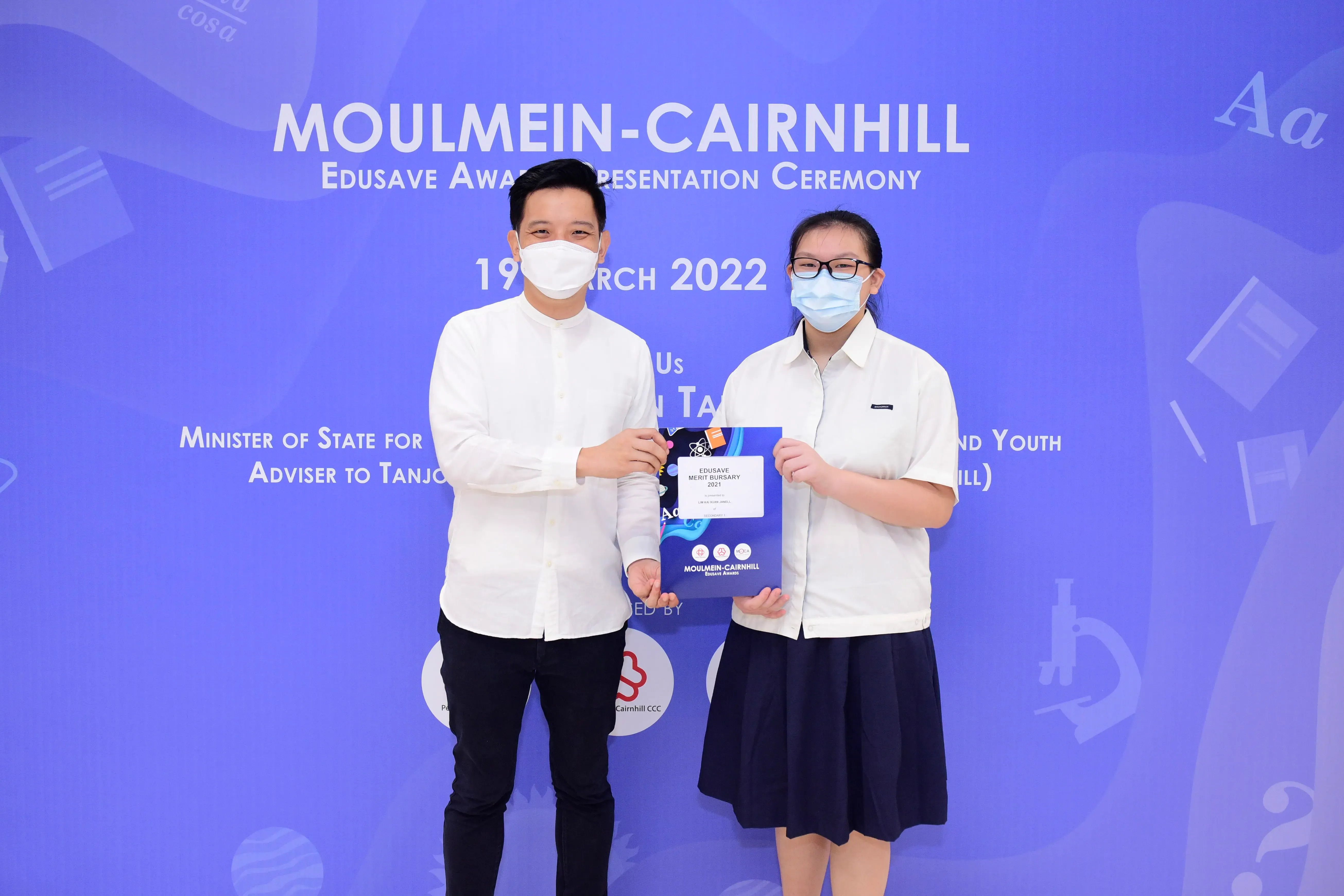 Two masked people hold an Edusave Merit Bursary certificate at the Moulmein-Cairnhill Award Ceremony, March 2022.