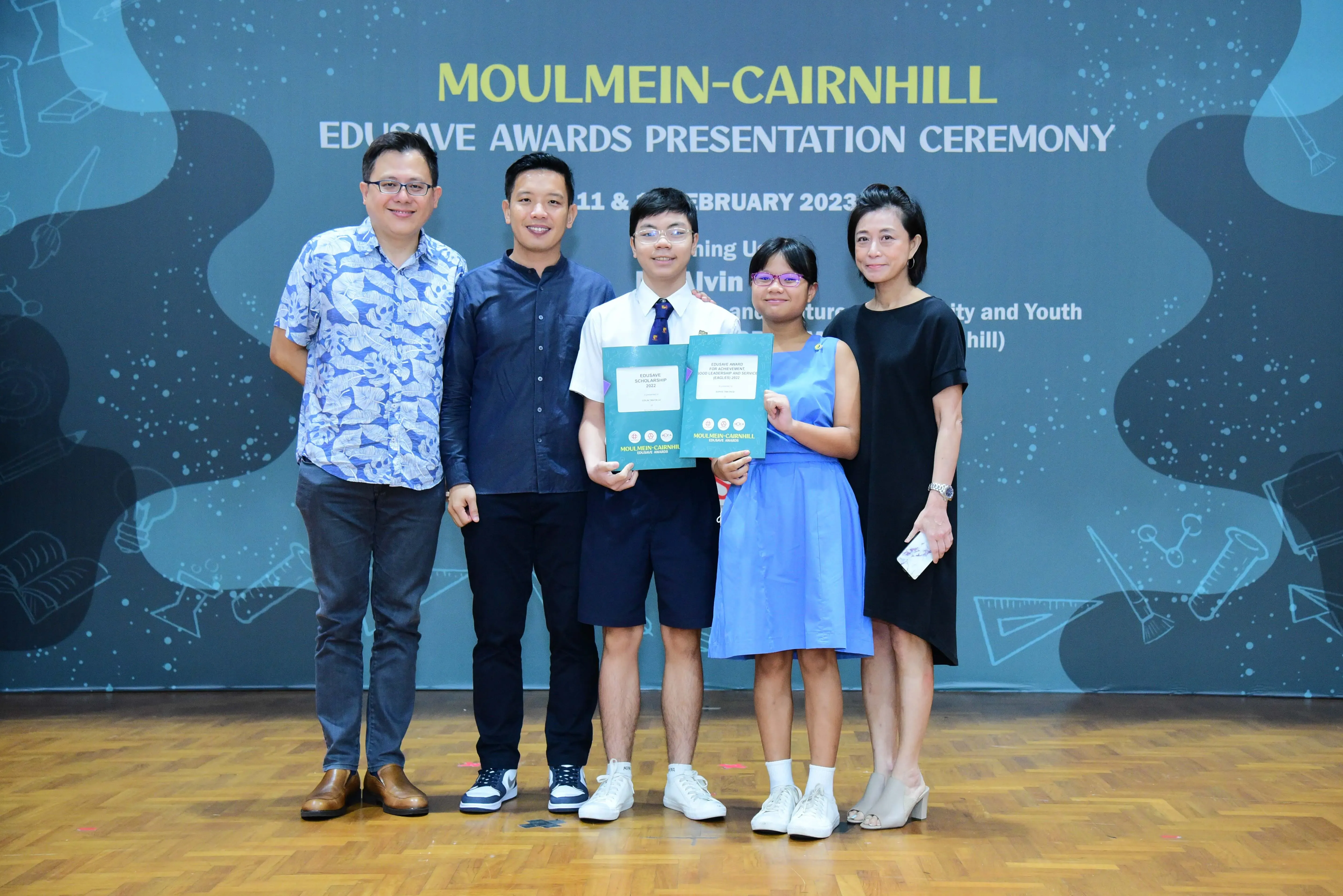 Five people pose at the Moulmein-Cairnhill Edusave Awards Presentation Ceremony, with two students holding green award certificates.
