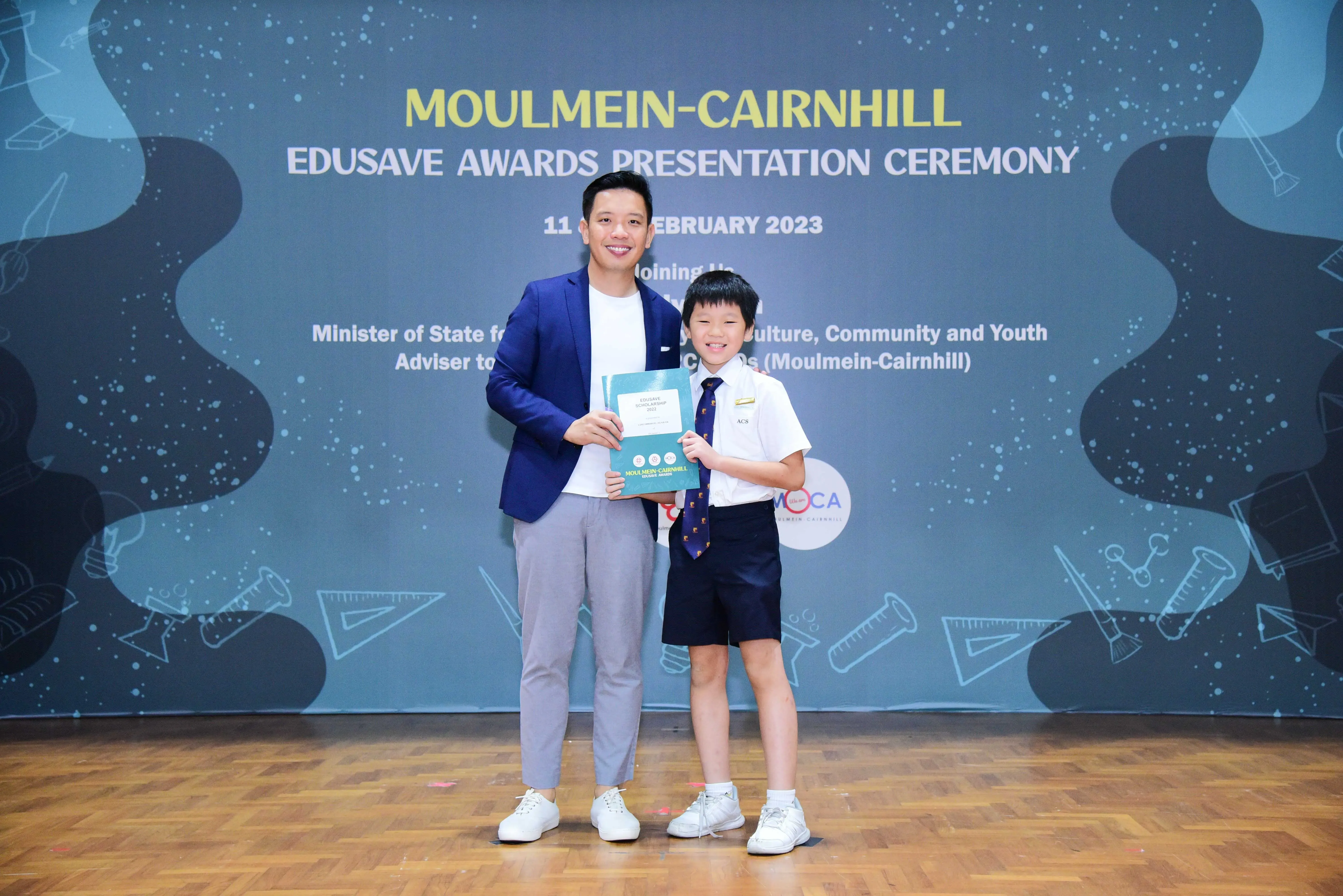 A man in a blue blazer and a young boy in school uniform hold a certificate at the Moulmein-Cairnhill Edusave Awards 2023.