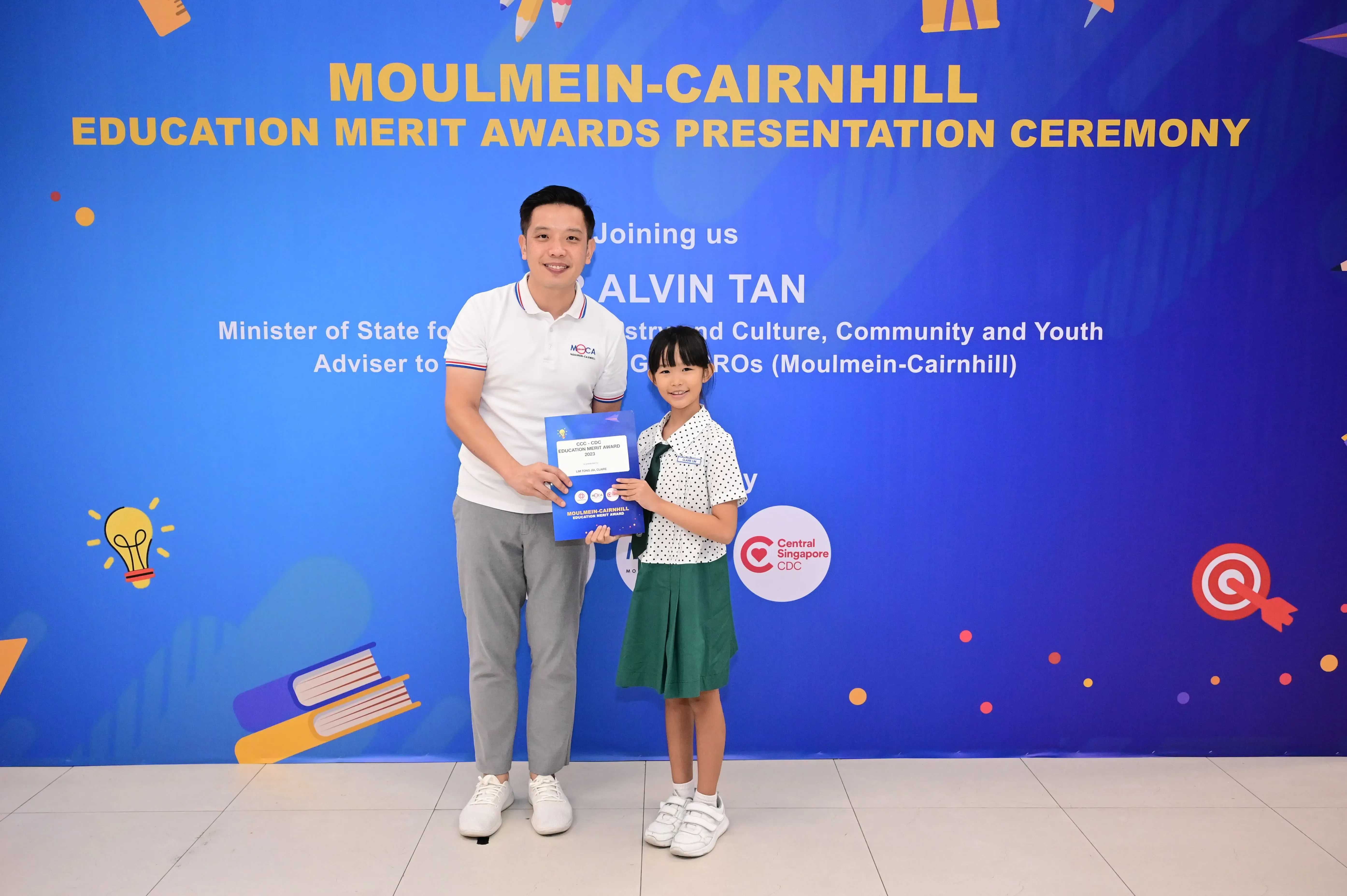 Man and young girl hold an award certificate in front of a Moulmein-Cairnhill Education Merit Awards banner.