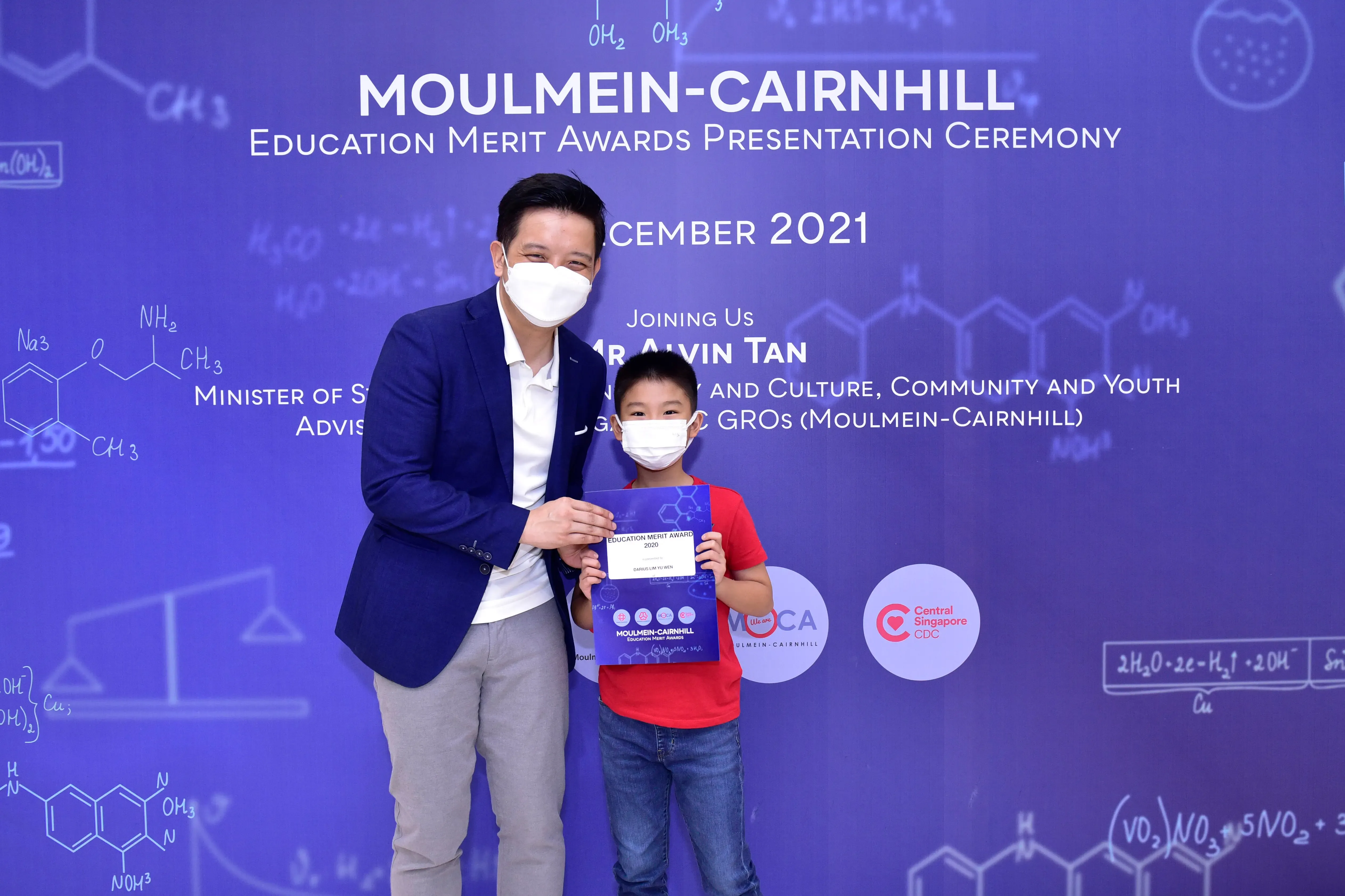 An adult and a young boy wearing masks hold an award certificate at the Moulmein-Cairnhill Education Merit Awards 2021.
