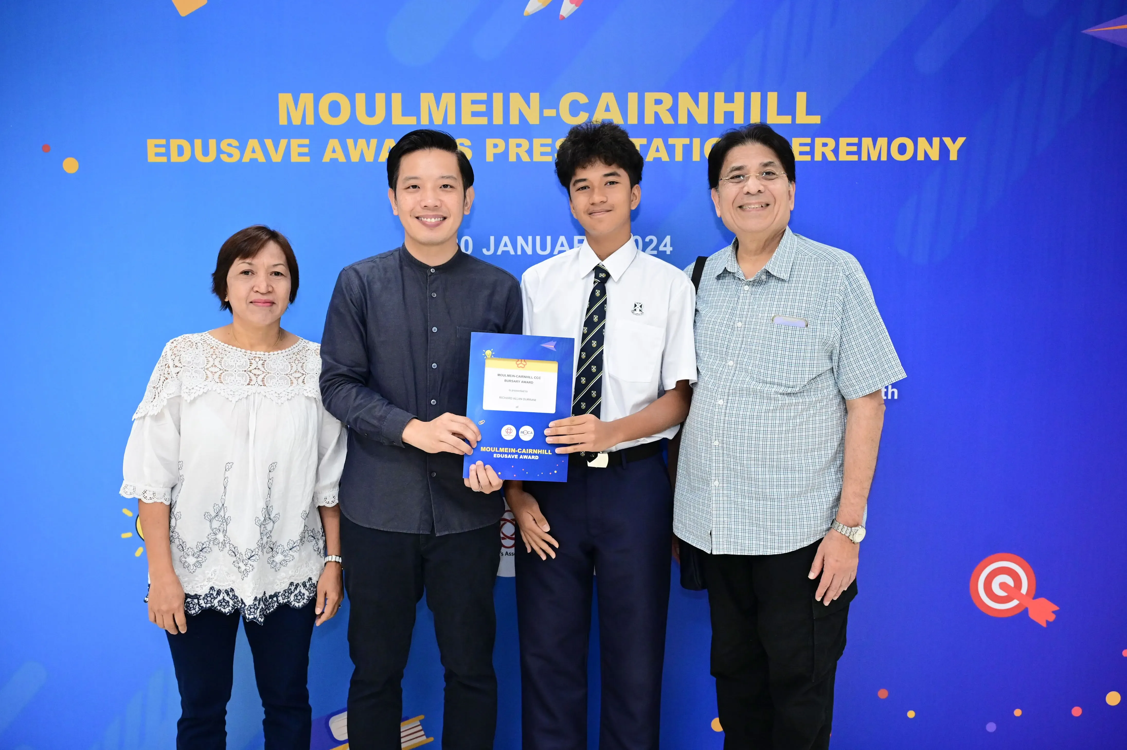 Four people smile at the Moulmein-Cairnhill Edusave Awards Presentation Ceremony, with a student holding a certificate.