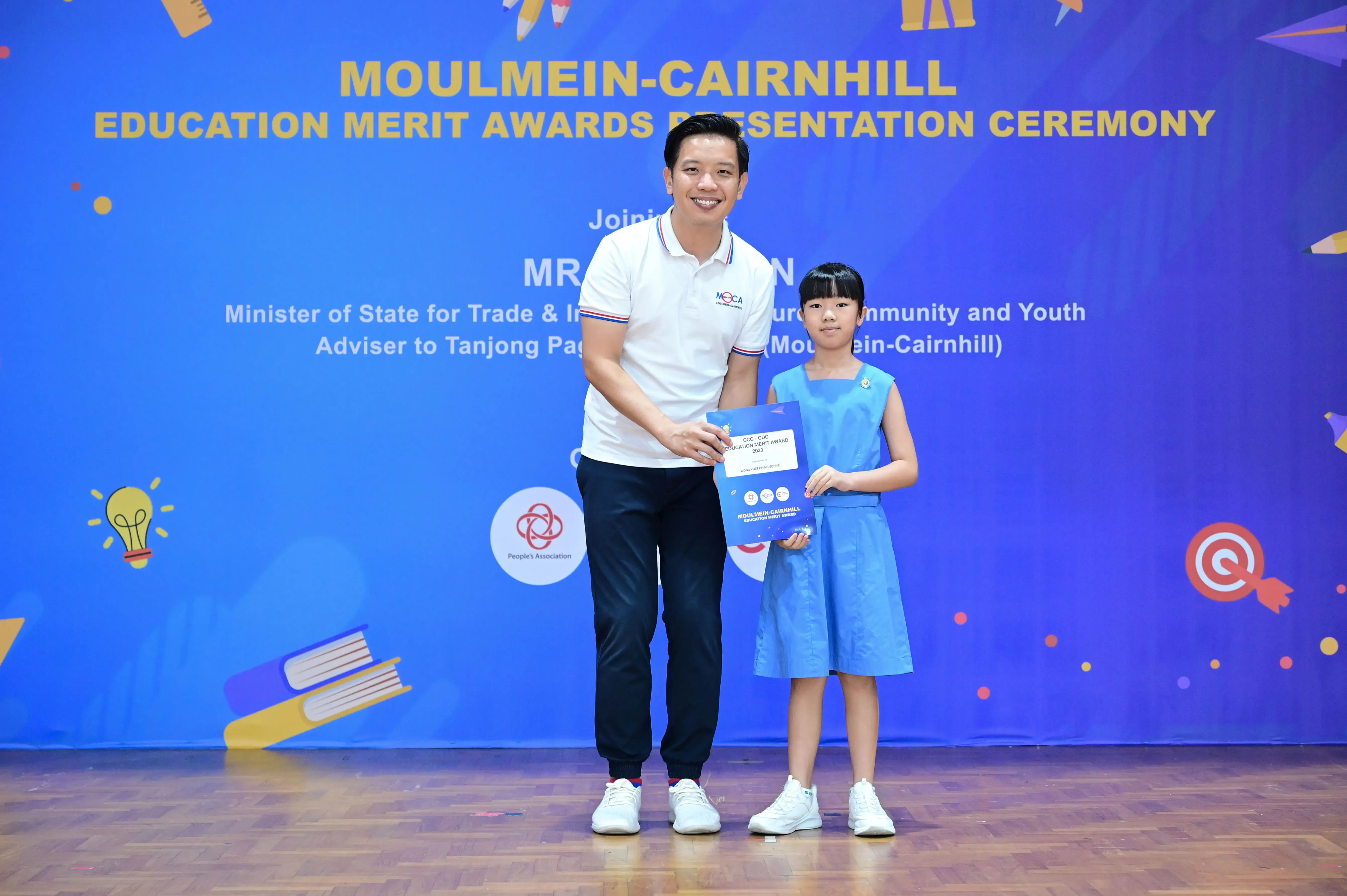 A man and a young girl hold an award certificate on stage at the Moulmein-Cairnhill Education Merit Awards ceremony.