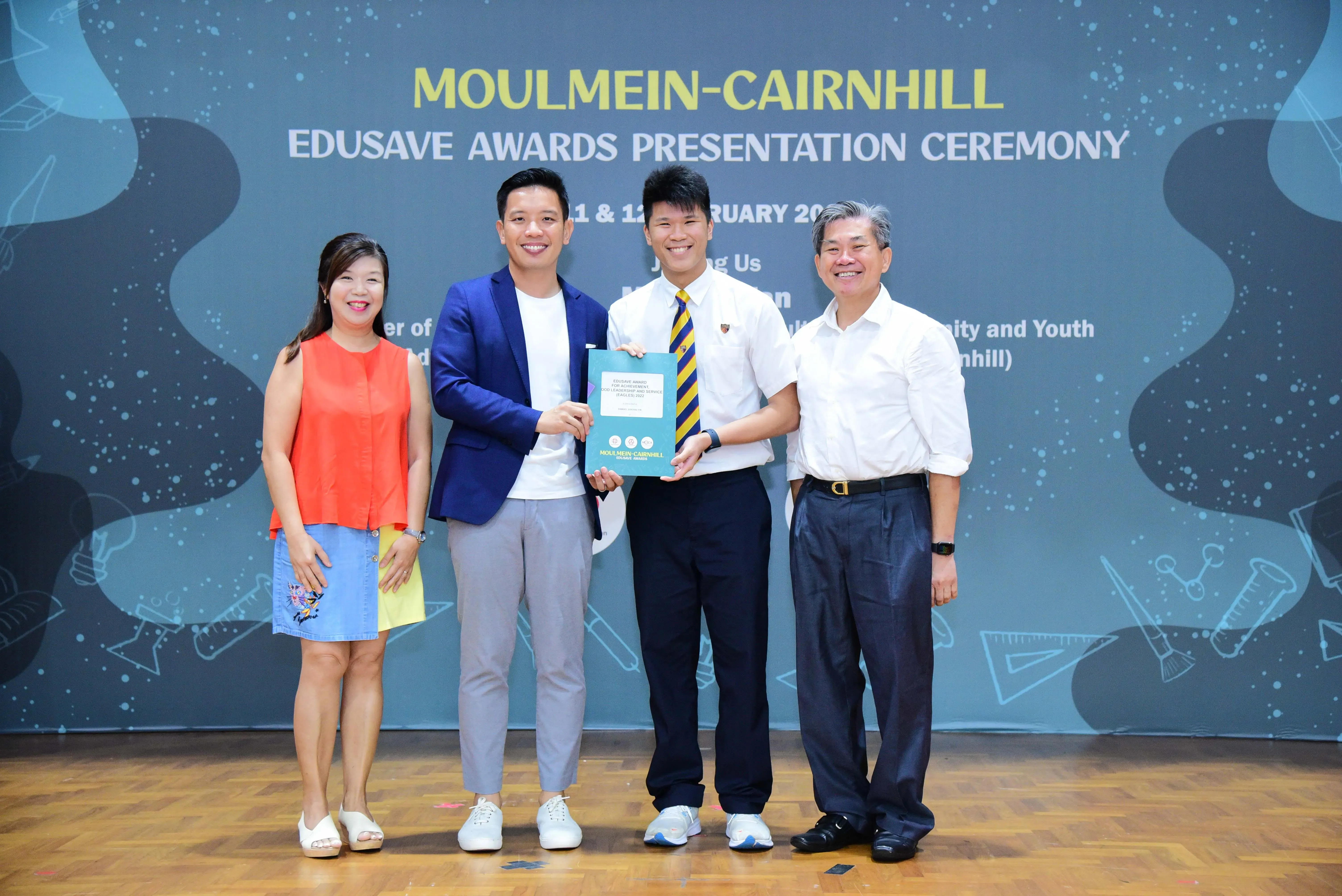 Four people smile at the Moulmein-Cairnhill Edusave Awards Presentation Ceremony as a student receives a certificate.
