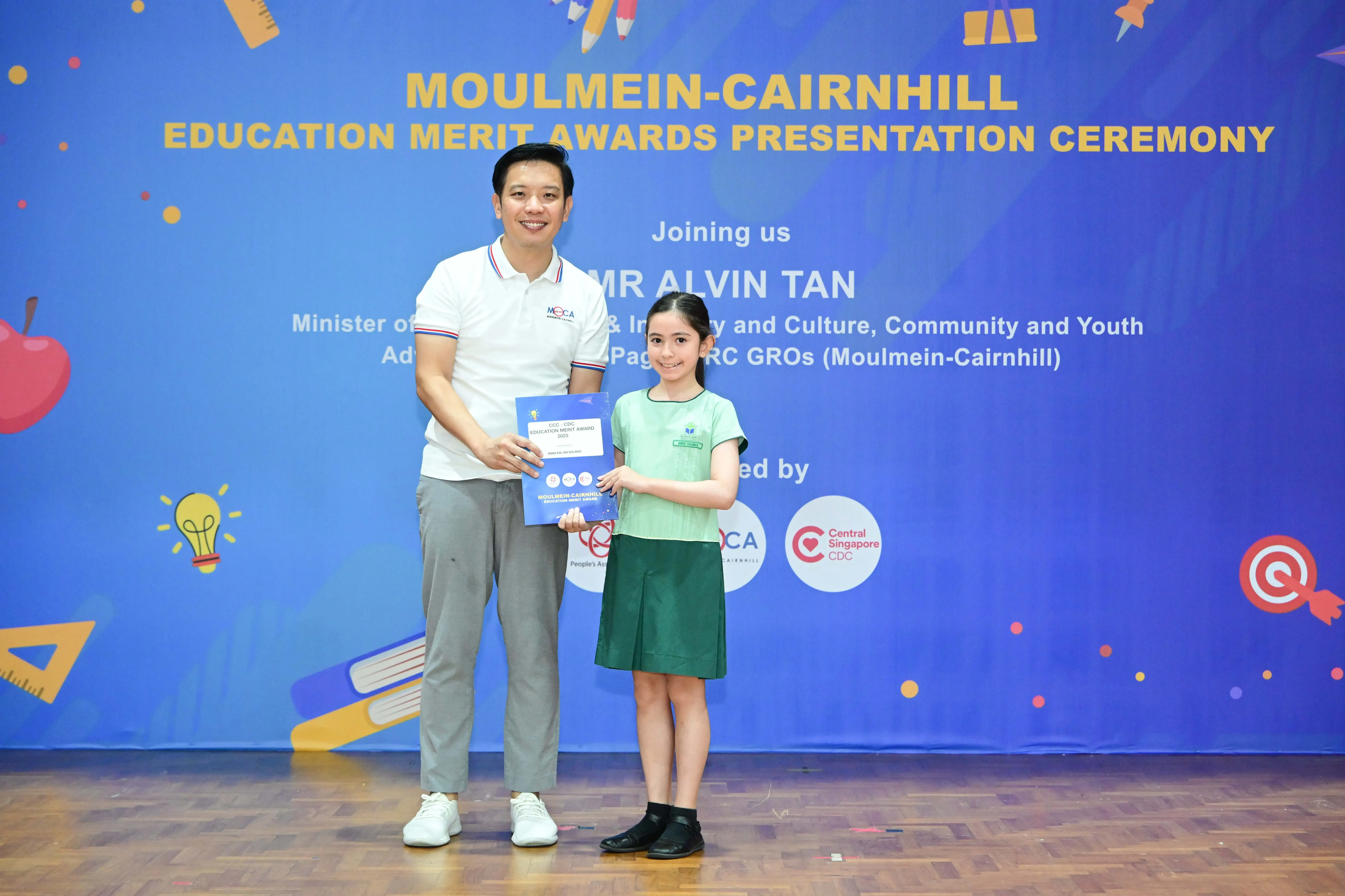 A man and a young girl hold a certificate in front of a Moulmein-Cairnhill Education Merit Awards banner.