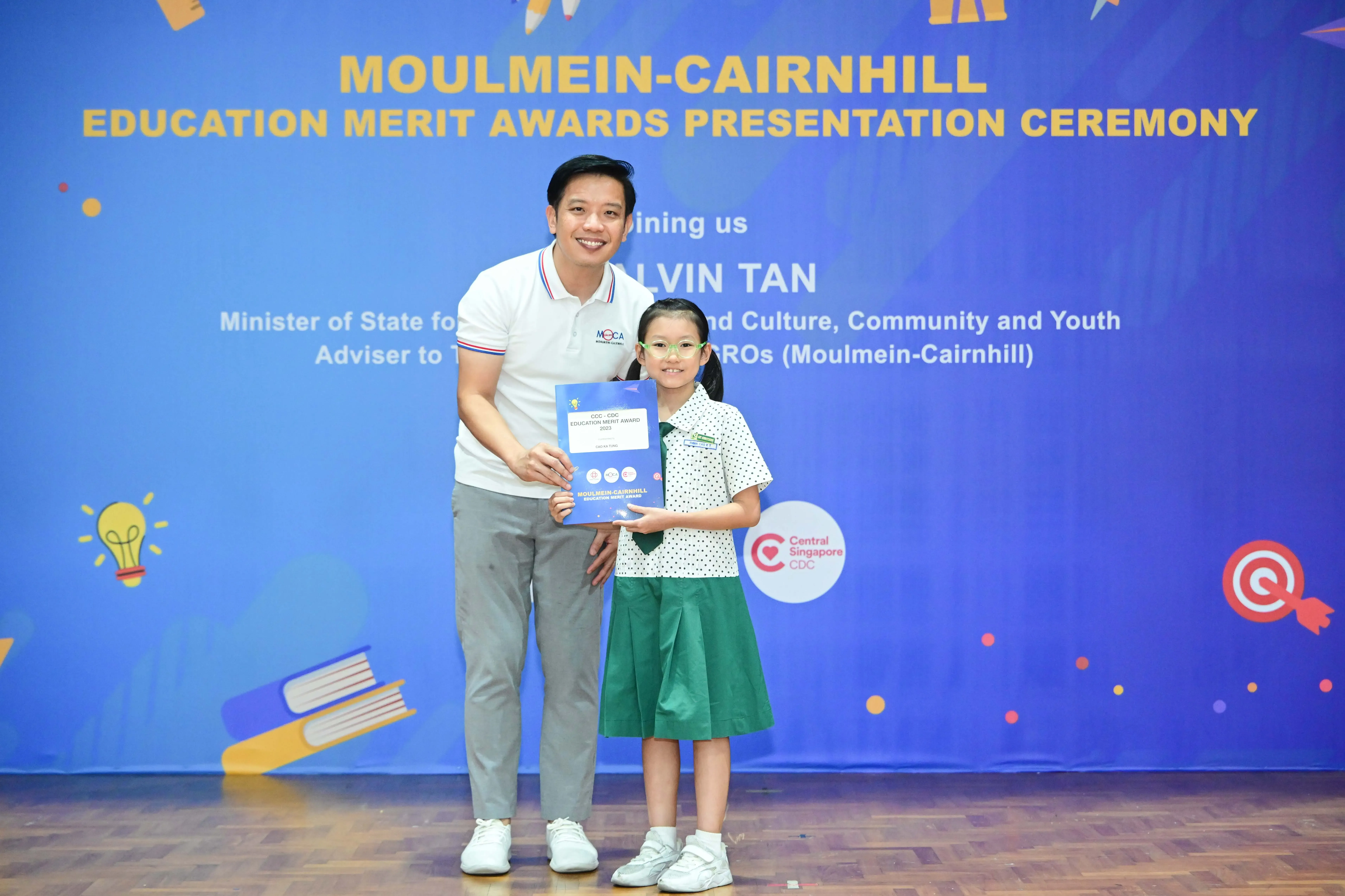 A man and a young girl hold a blue award certificate at the Moulmein-Cairnhill Education Merit Awards ceremony.