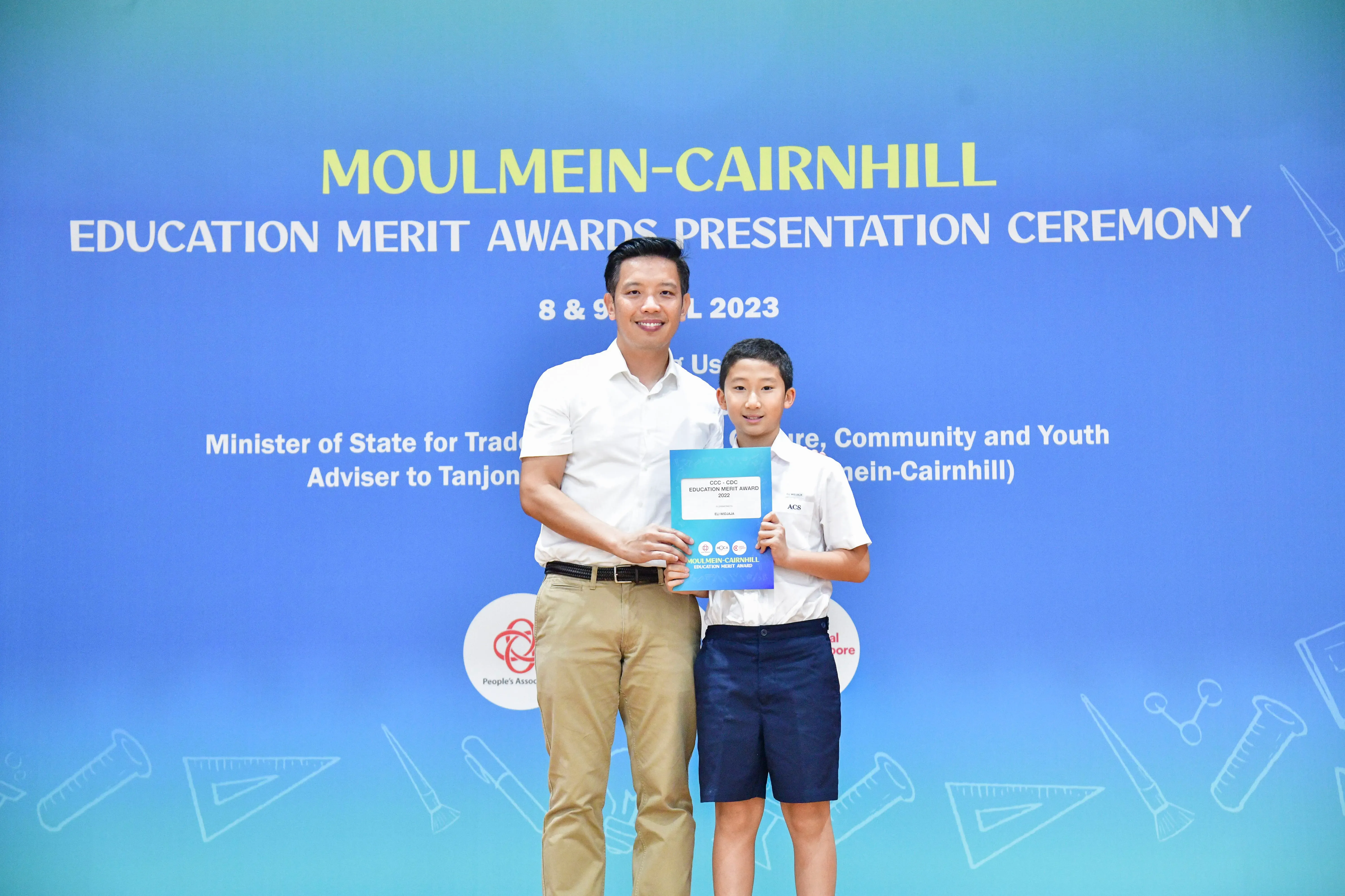 A man and a young boy hold an award certificate in front of a Moulmein-Cairnhill Education Merit Awards 2023 banner.