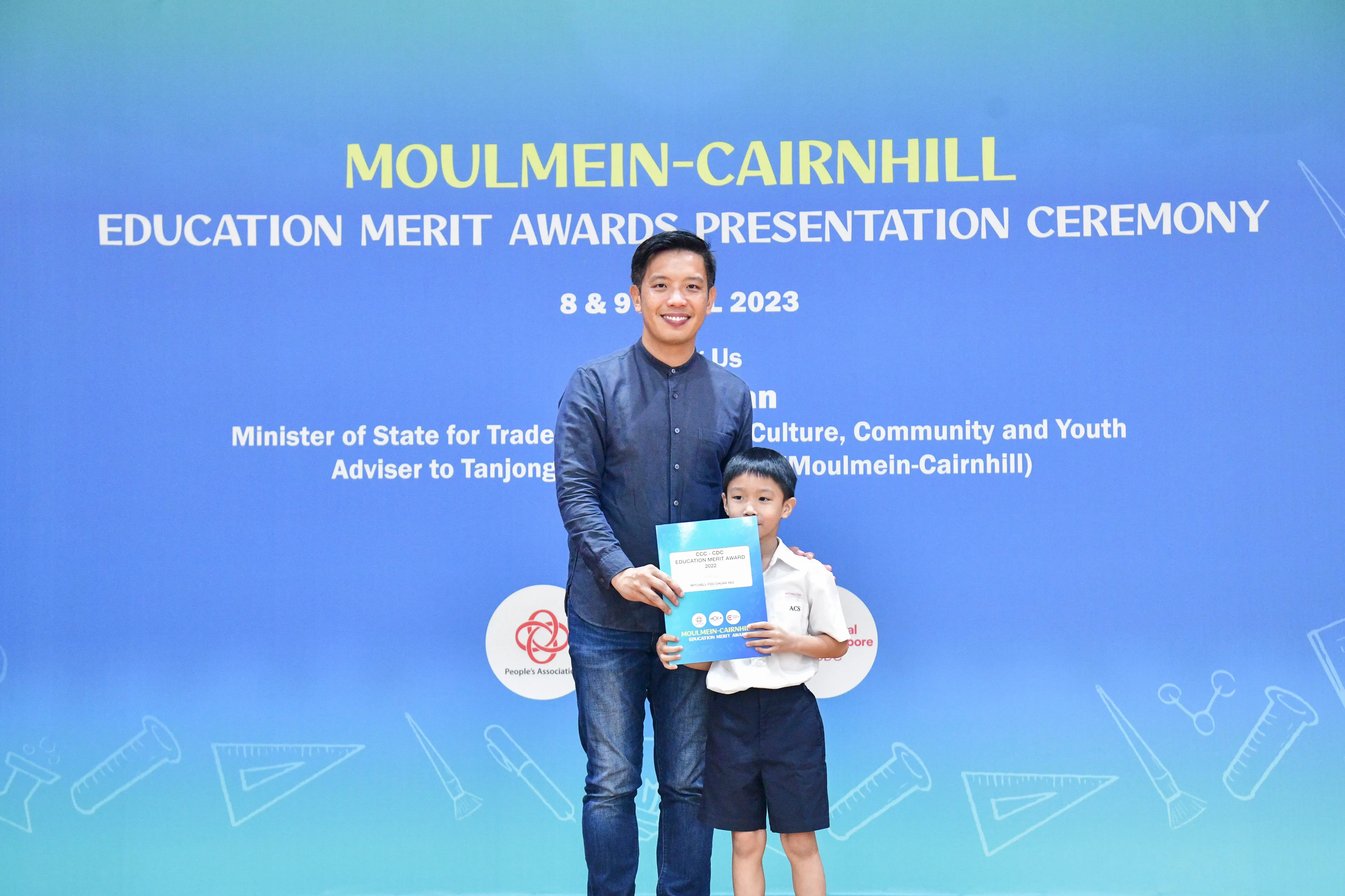 A man and a young boy hold a certificate in front of a Moulmein-Cairnhill Education Merit Awards 2023 banner.