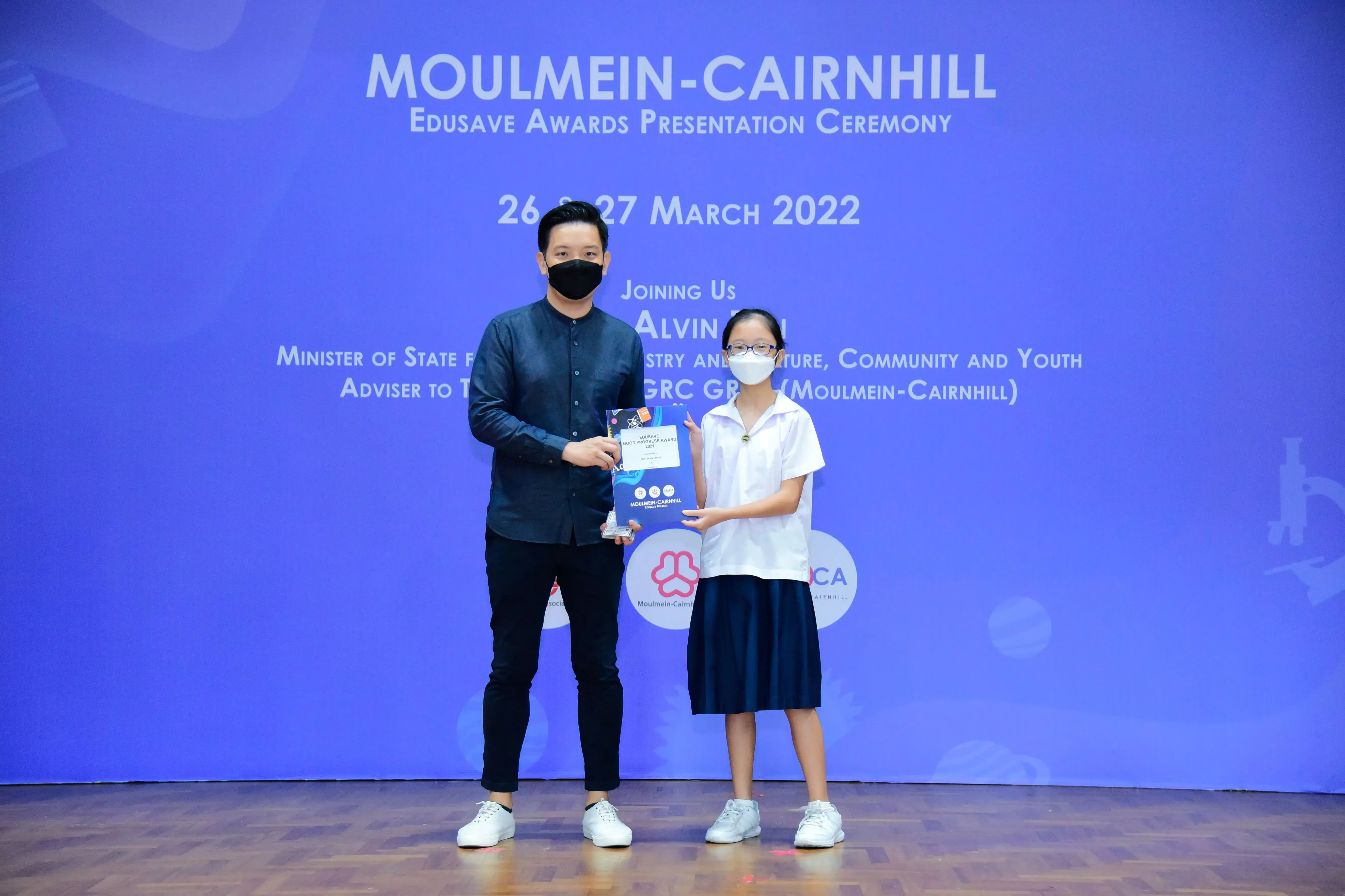 A masked man and a young girl in school uniform hold an award at the Moulmein-Cairnhill Edusave Awards Ceremony 2022.
