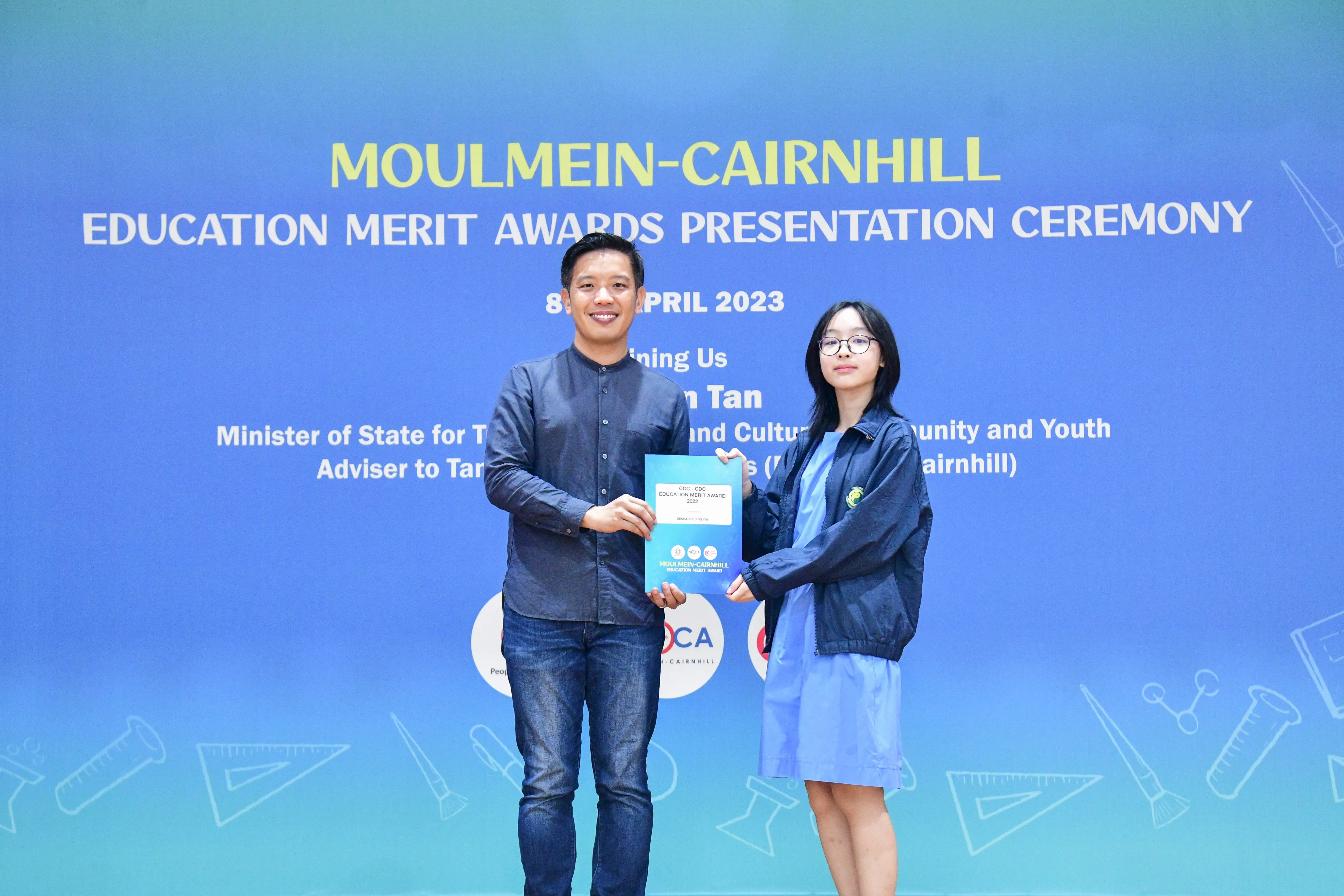 Two people holding a certificate in front of a blue Moulmein-Cairnhill Education Merit Awards banner dated April 2023.