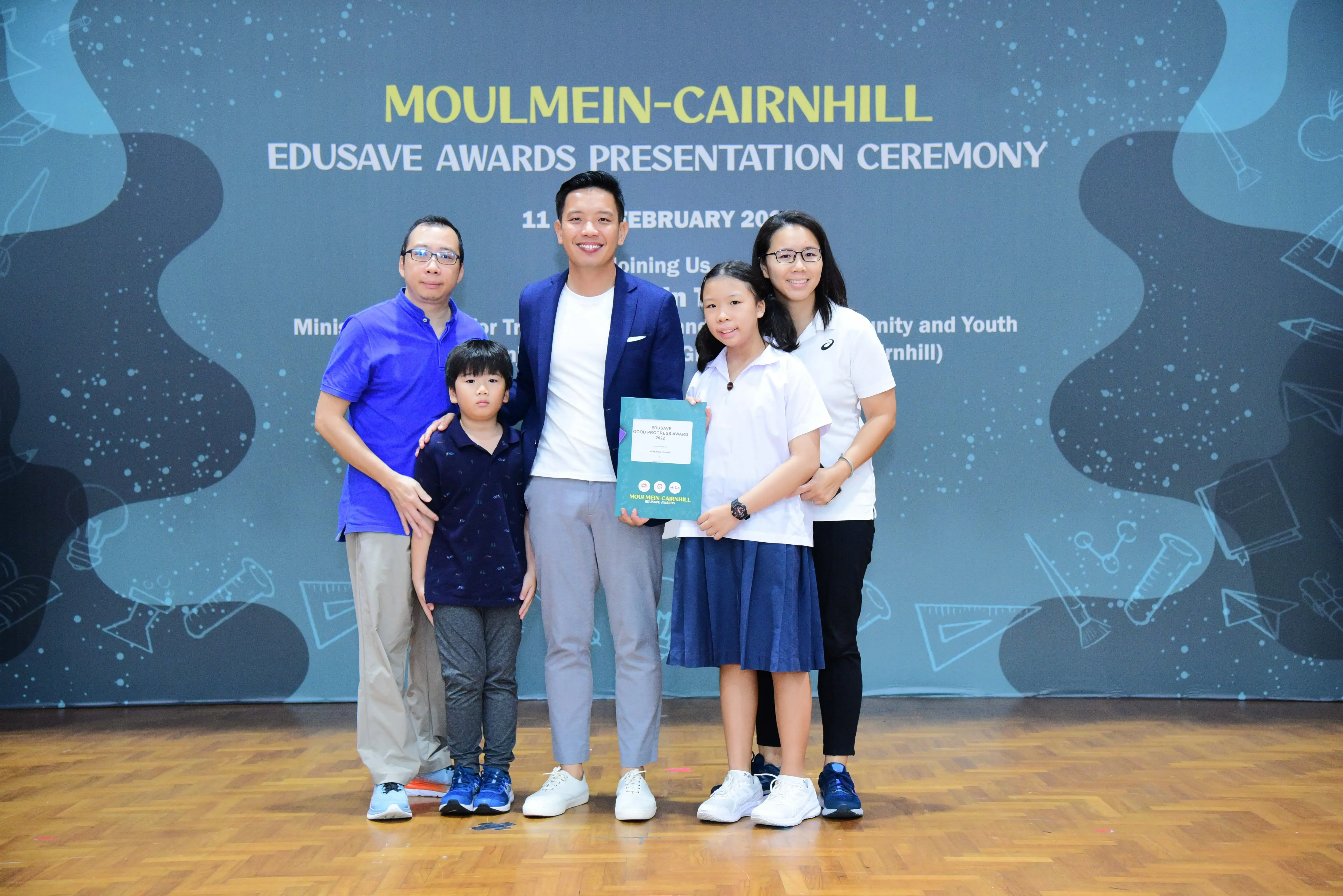 Five people pose at the Moulmein-Cairnhill Edusave Awards Presentation Ceremony, including a girl holding an award folder.