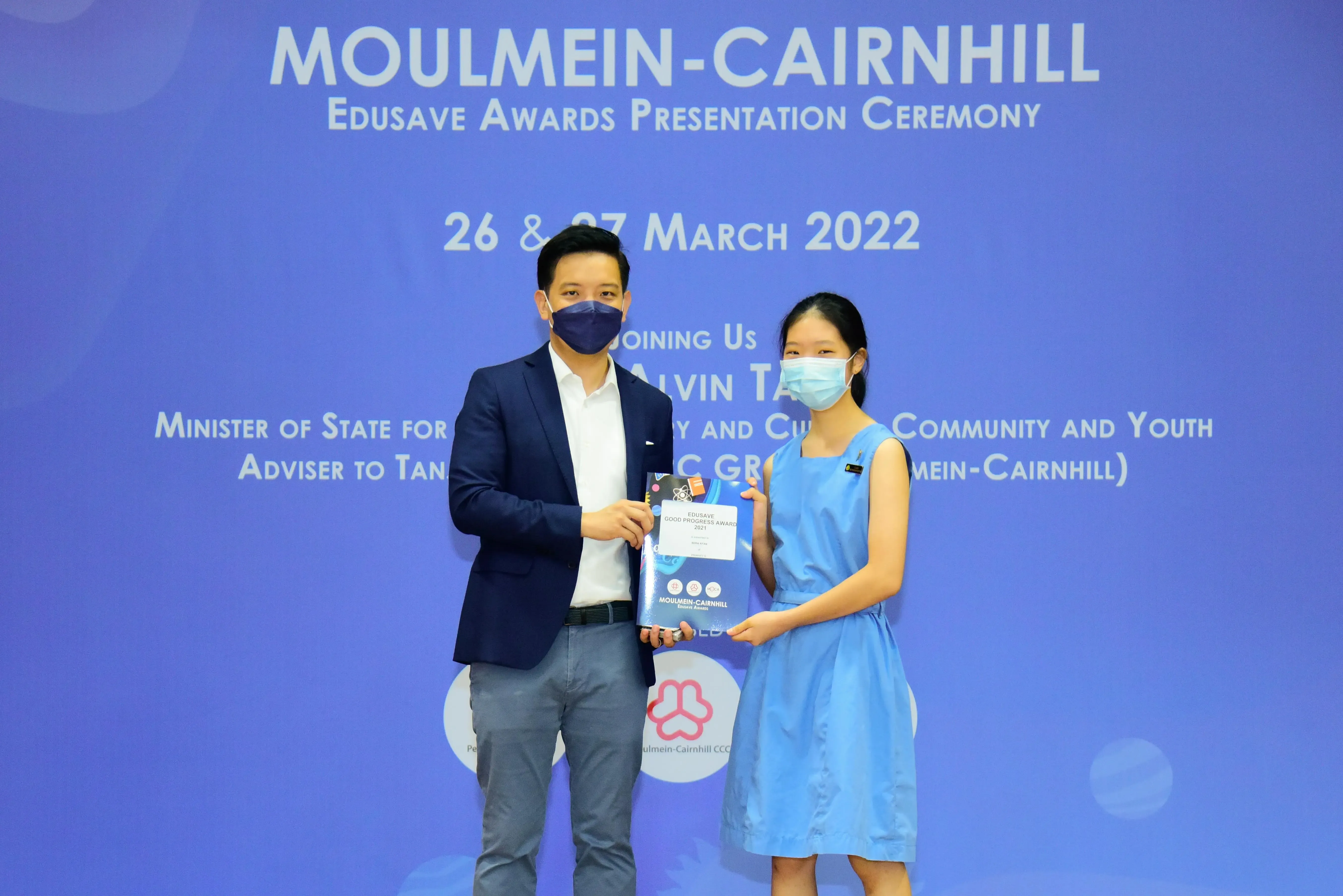 Two masked people hold an Edusave award at the Moulmein-Cairnhill Edusave Awards Presentation Ceremony, March 2022.