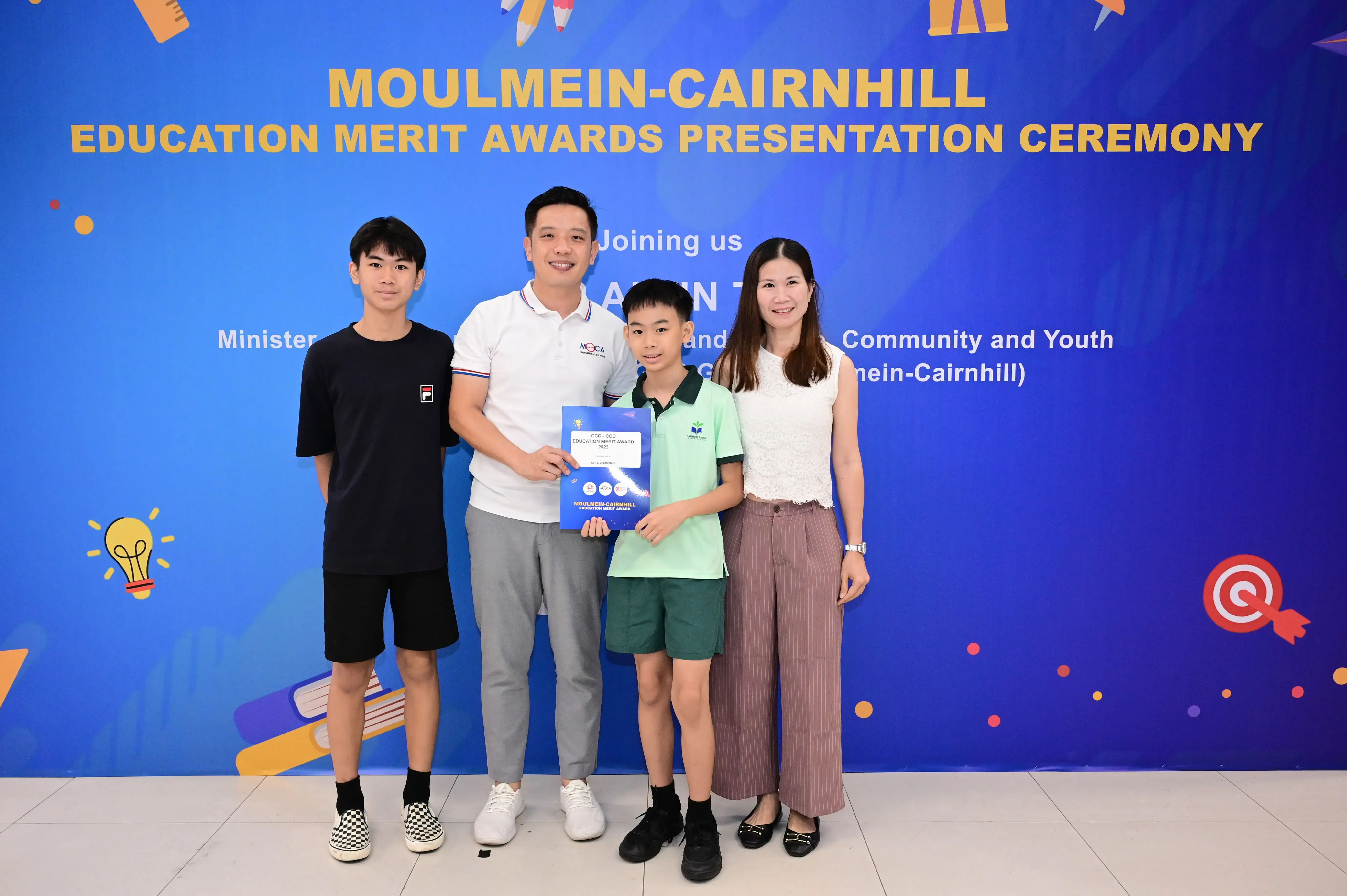 Four people pose in front of a Moulmein-Cairnhill Education Merit Awards Presentation Ceremony banner, with a boy holding a certificate.