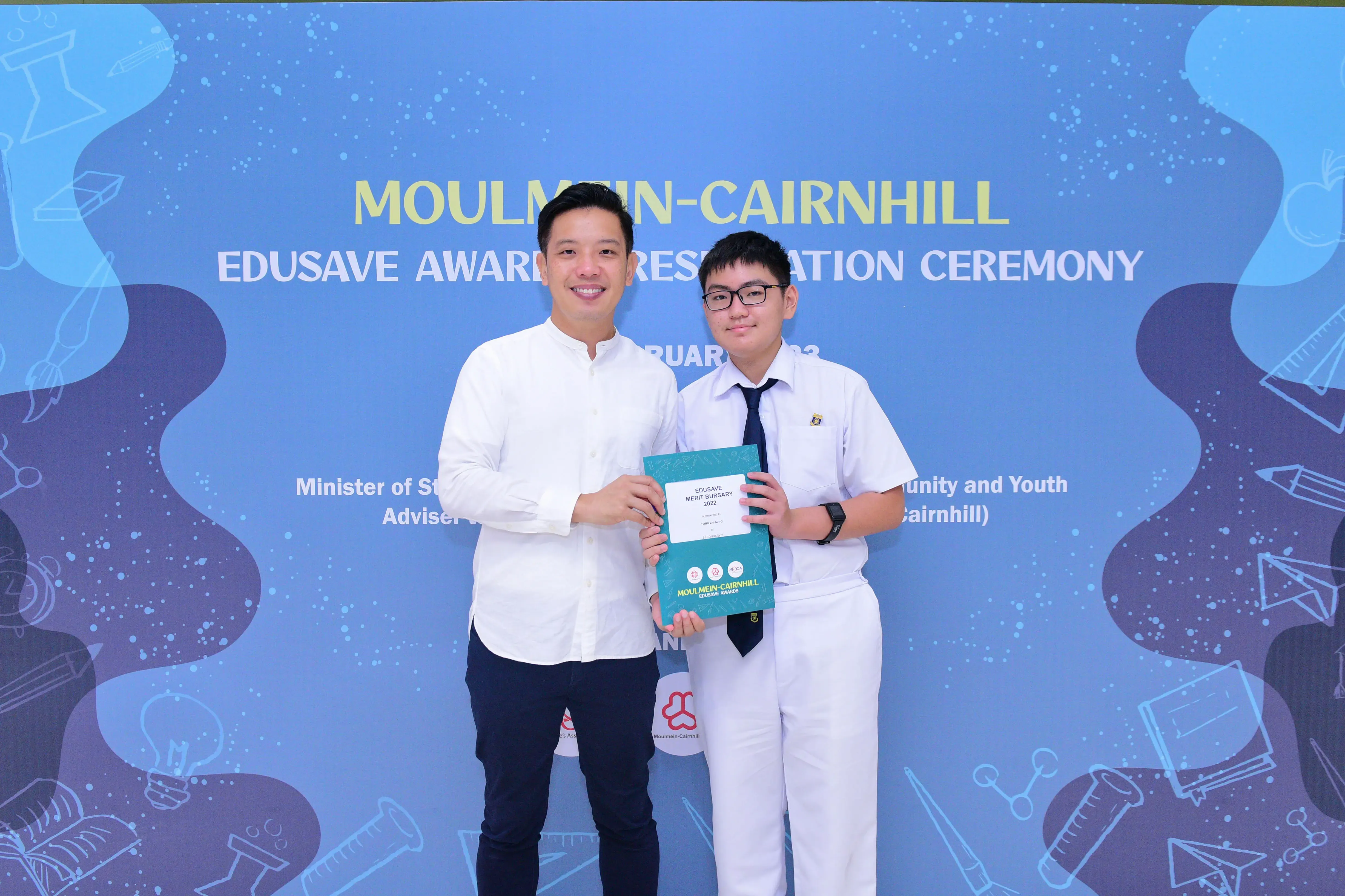 Two people hold a teal award folder at the Moulmein-Cairnhill Edusave Awards Presentation Ceremony backdrop.