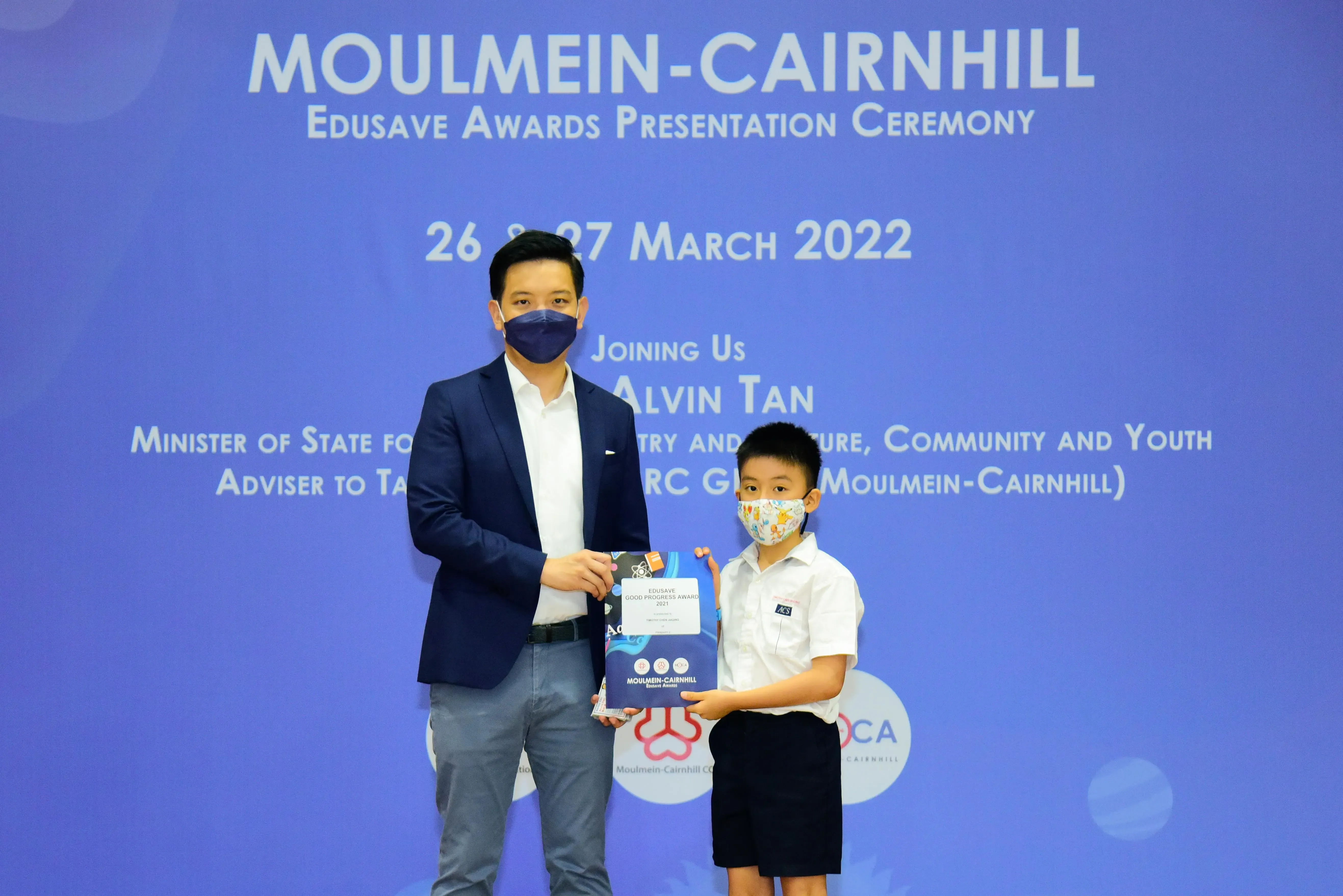 An adult in a suit and a young boy in school uniform hold an award at the Moulmein-Cairnhill Edusave Awards Ceremony.