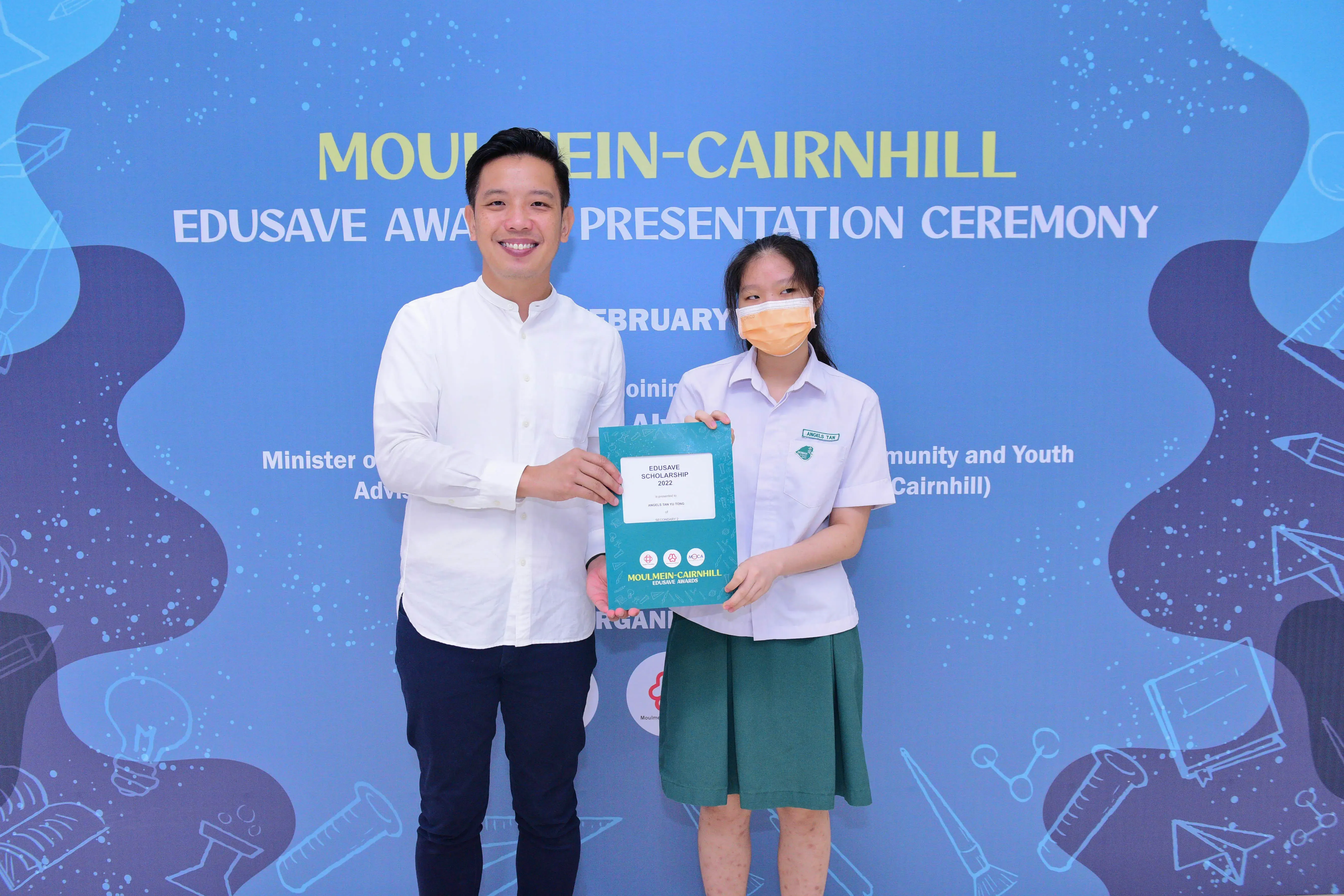 A man and a masked schoolgirl hold an Edusave Scholarship certificate at the Moulmein-Cairnhill Awards Ceremony.