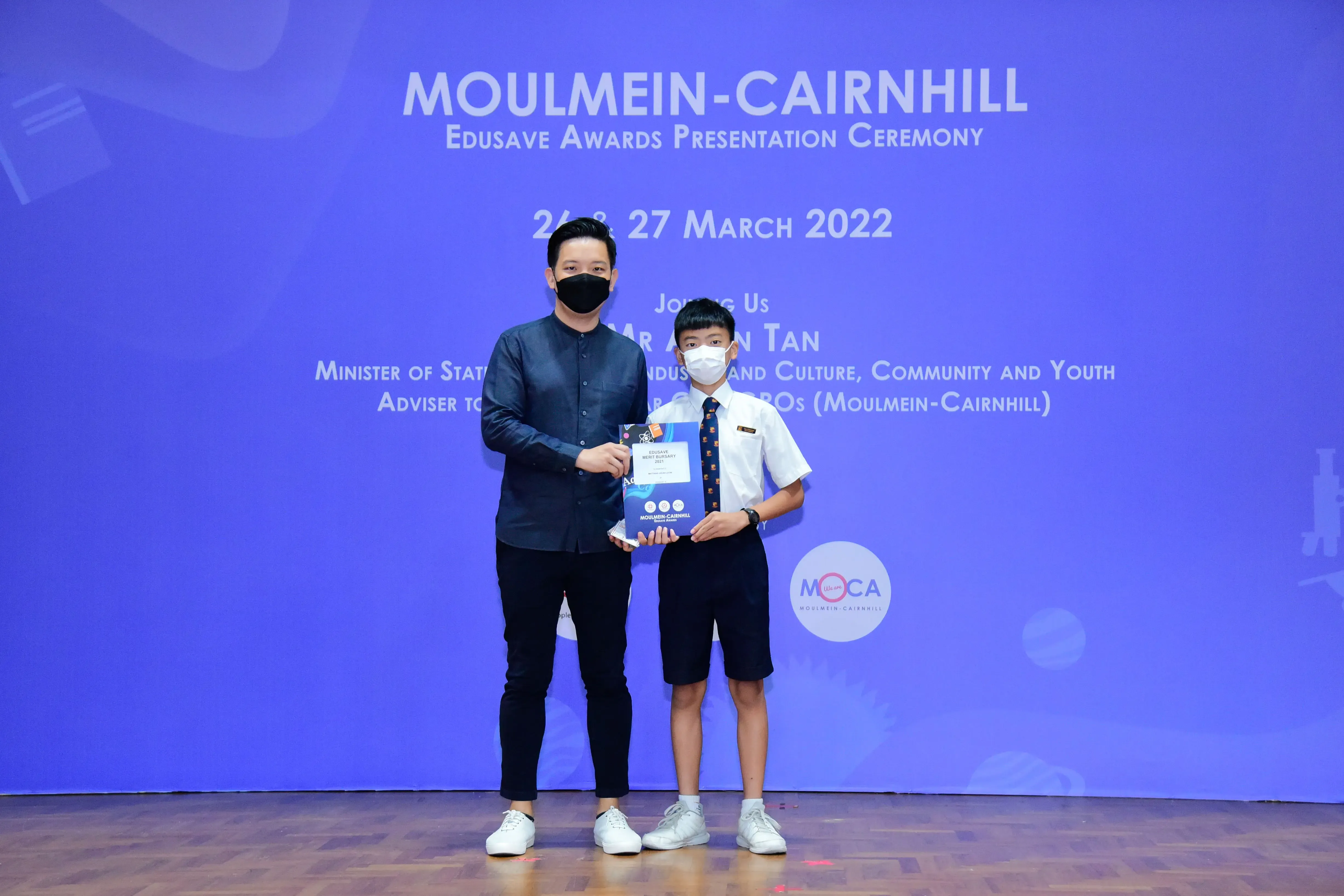 An adult and a masked student hold an award together at the Moulmein-Cairnhill Edusave Awards Ceremony 2022.