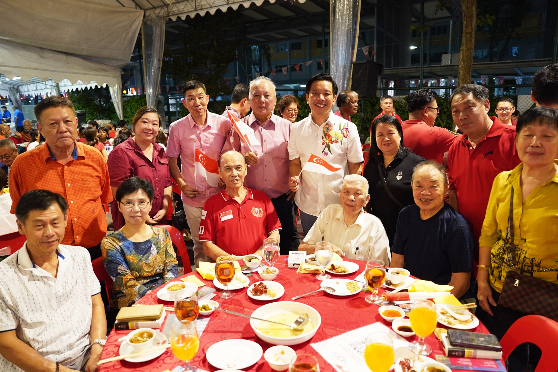 A large group of people posing at a red-themed outdoor dinner event, with Singapore flags and food on the table.