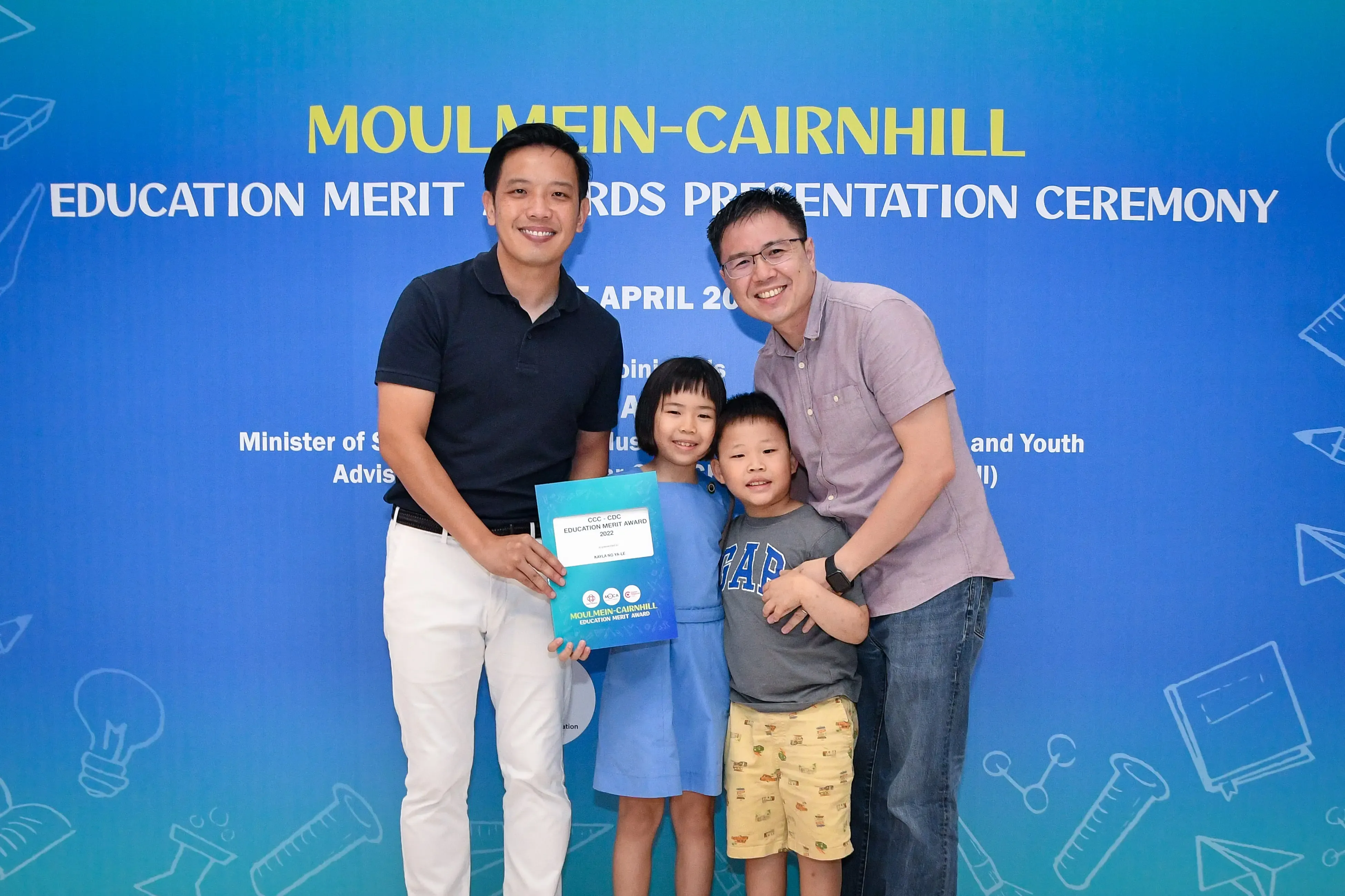 Two men and two children pose smiling at the Moulmein-Cairnhill Education Merit Awards Presentation Ceremony, holding an award certificate.