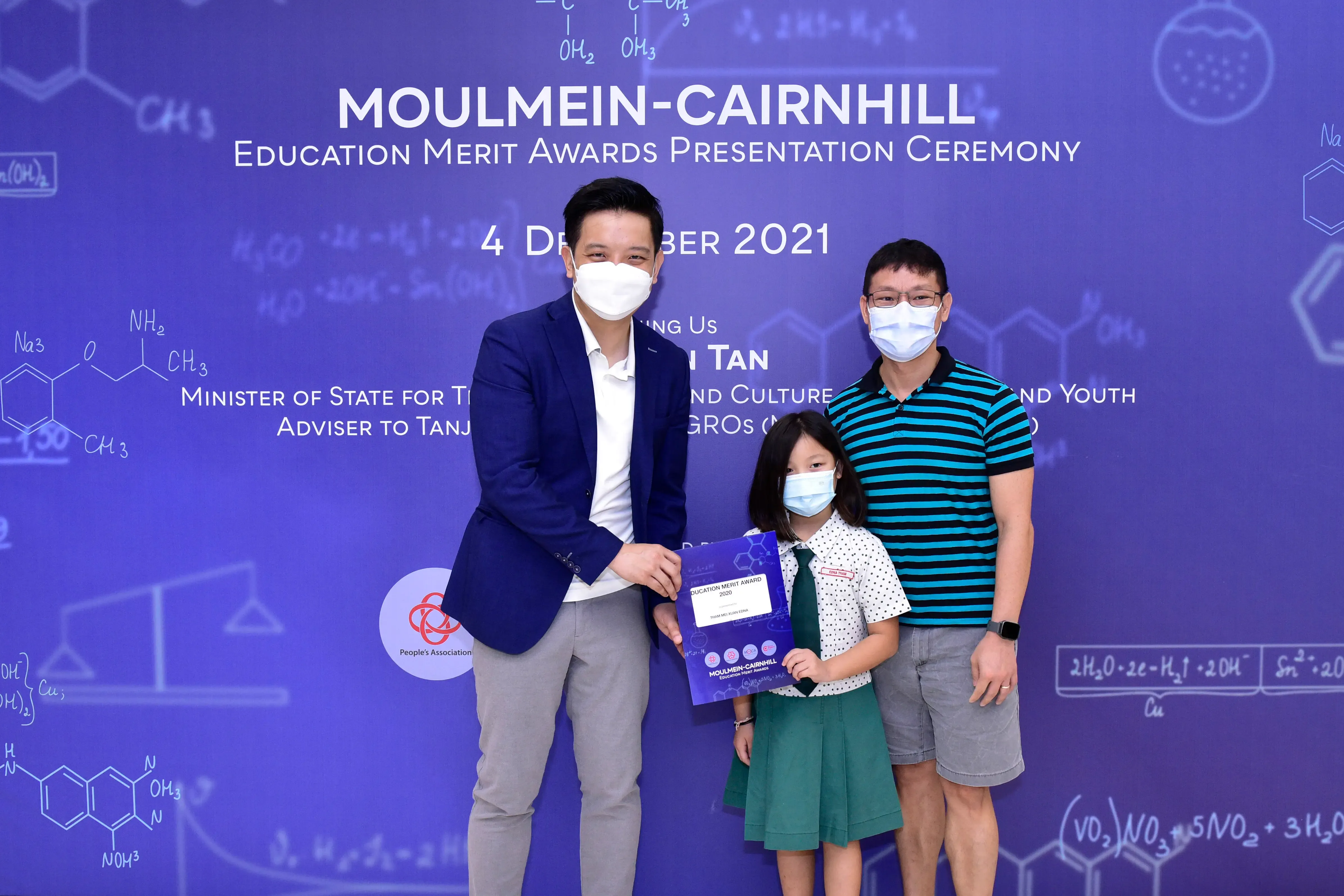 Two masked adults and a masked girl holding an award certificate at the Moulmein-Cairnhill Education Merit Awards 2021.