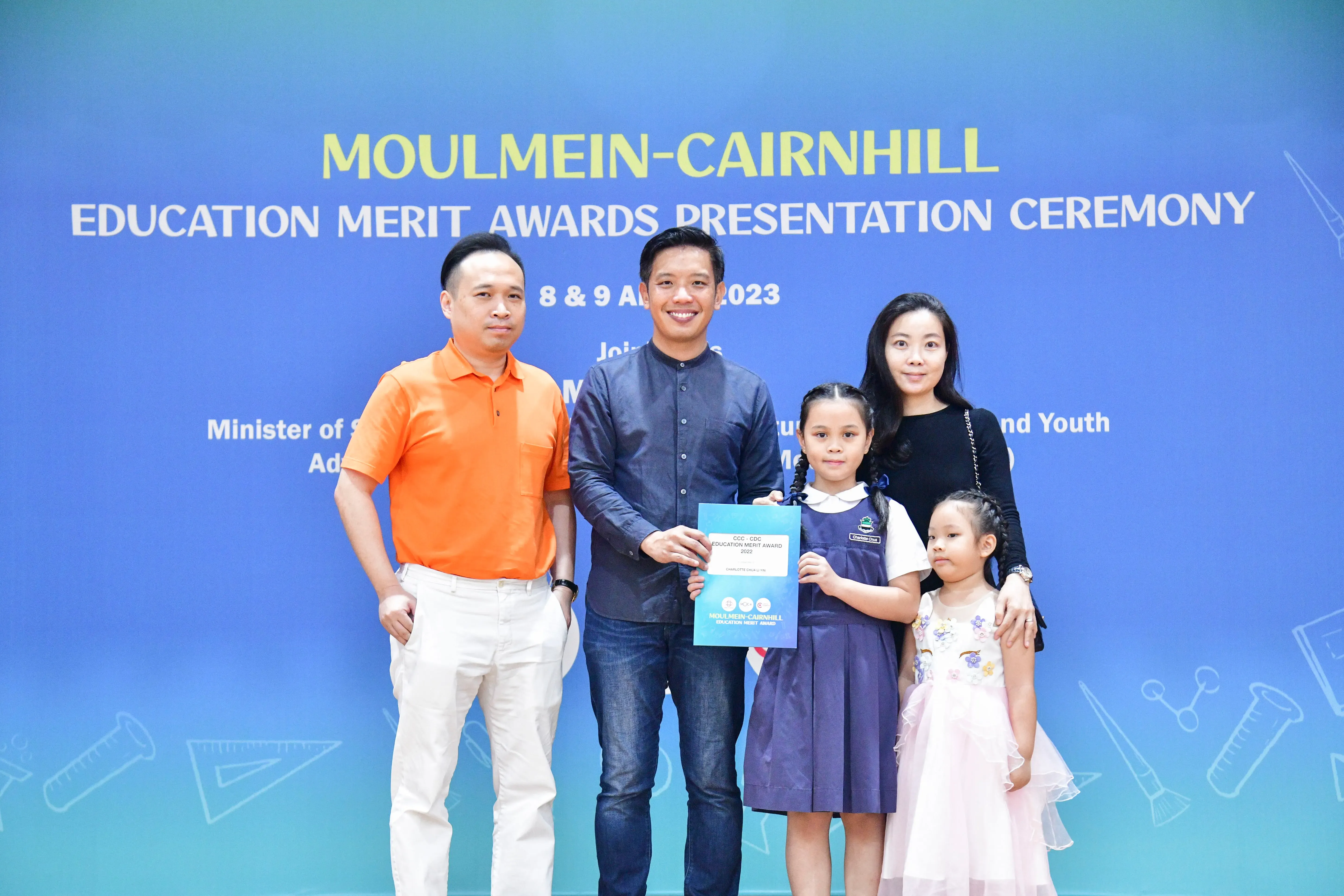 Five people pose before a Moulmein-Cairnhill Education Merit Awards Presentation Ceremony backdrop, with a girl holding a certificate.