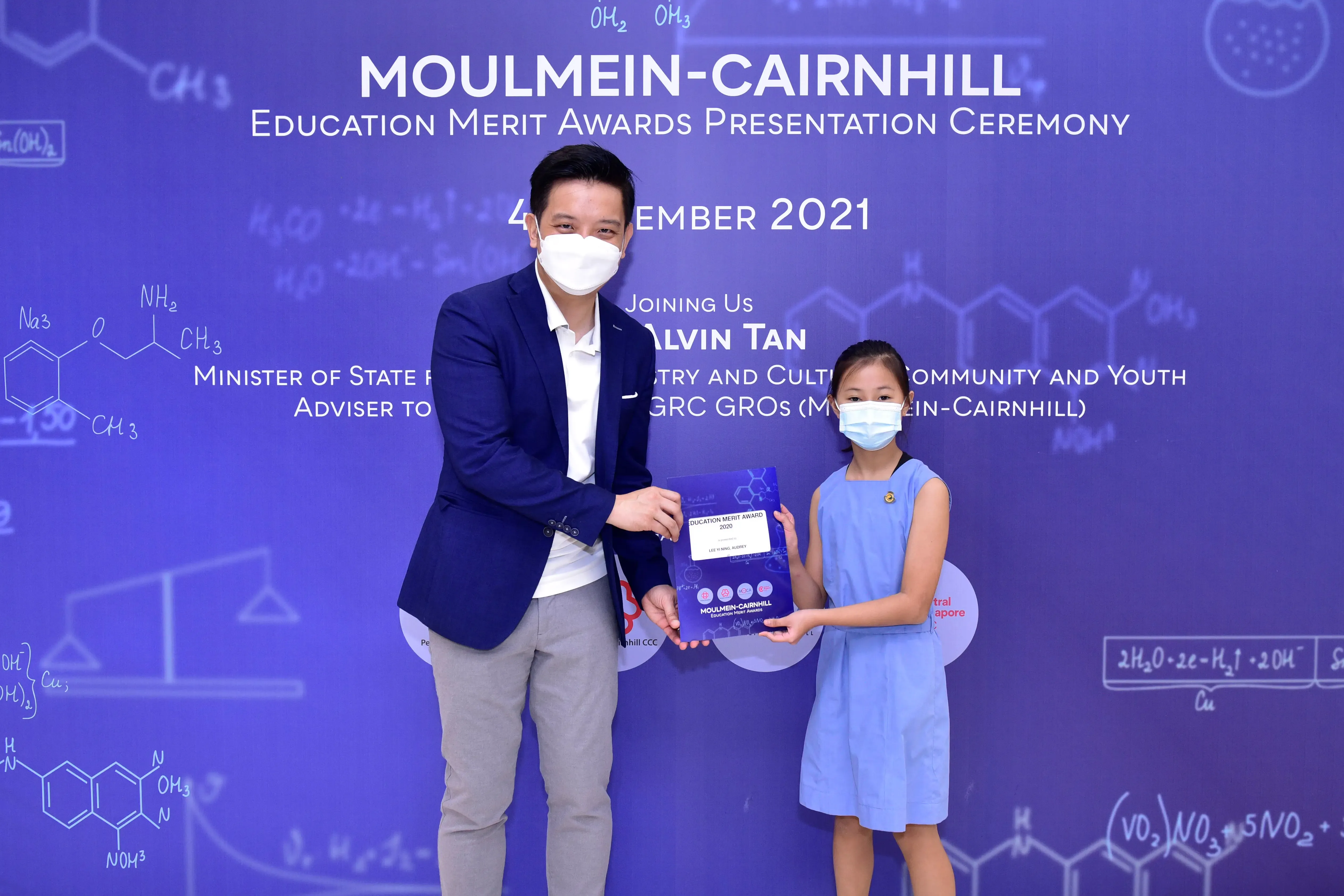A masked man in a blue blazer presents an award booklet to a young masked girl at the Moulmein-Cairnhill Education Merit Awards 2021.