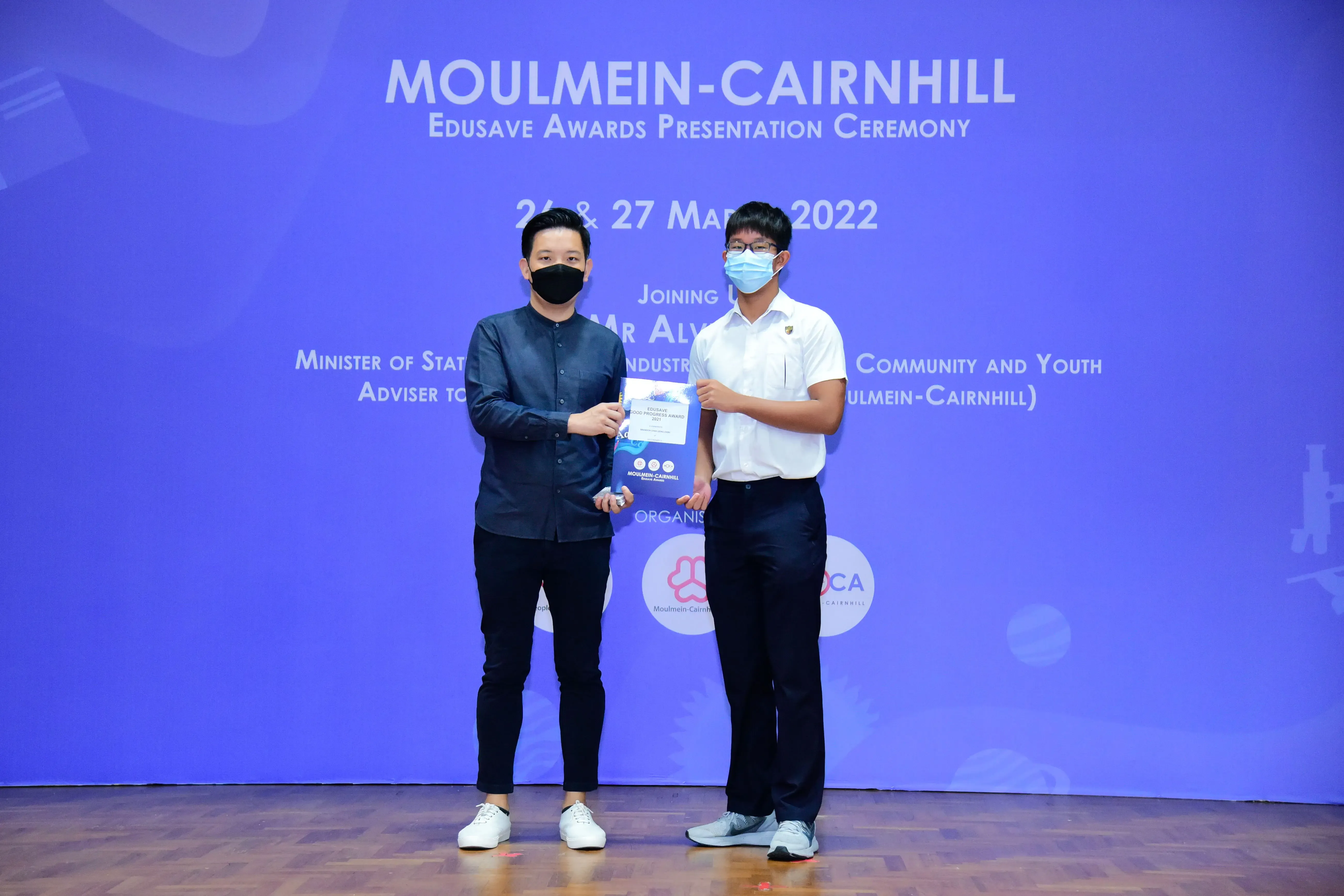 Two masked men hold a certificate on stage at the Moulmein-Cairnhill Edusave Awards Ceremony, March 2022.