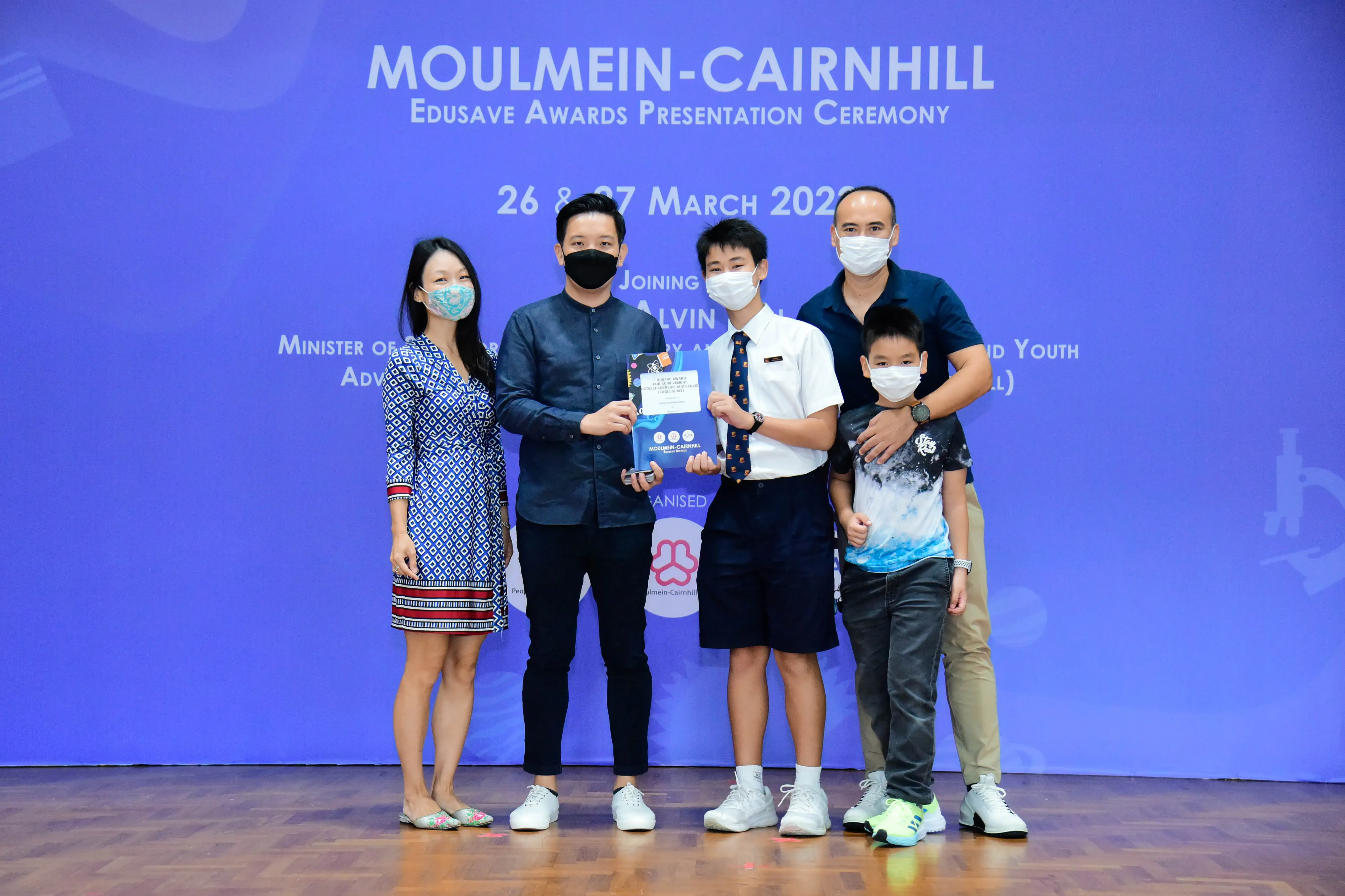Five masked people pose on stage at the Moulmein-Cairnhill Edusave Awards Presentation Ceremony, holding a trophy.