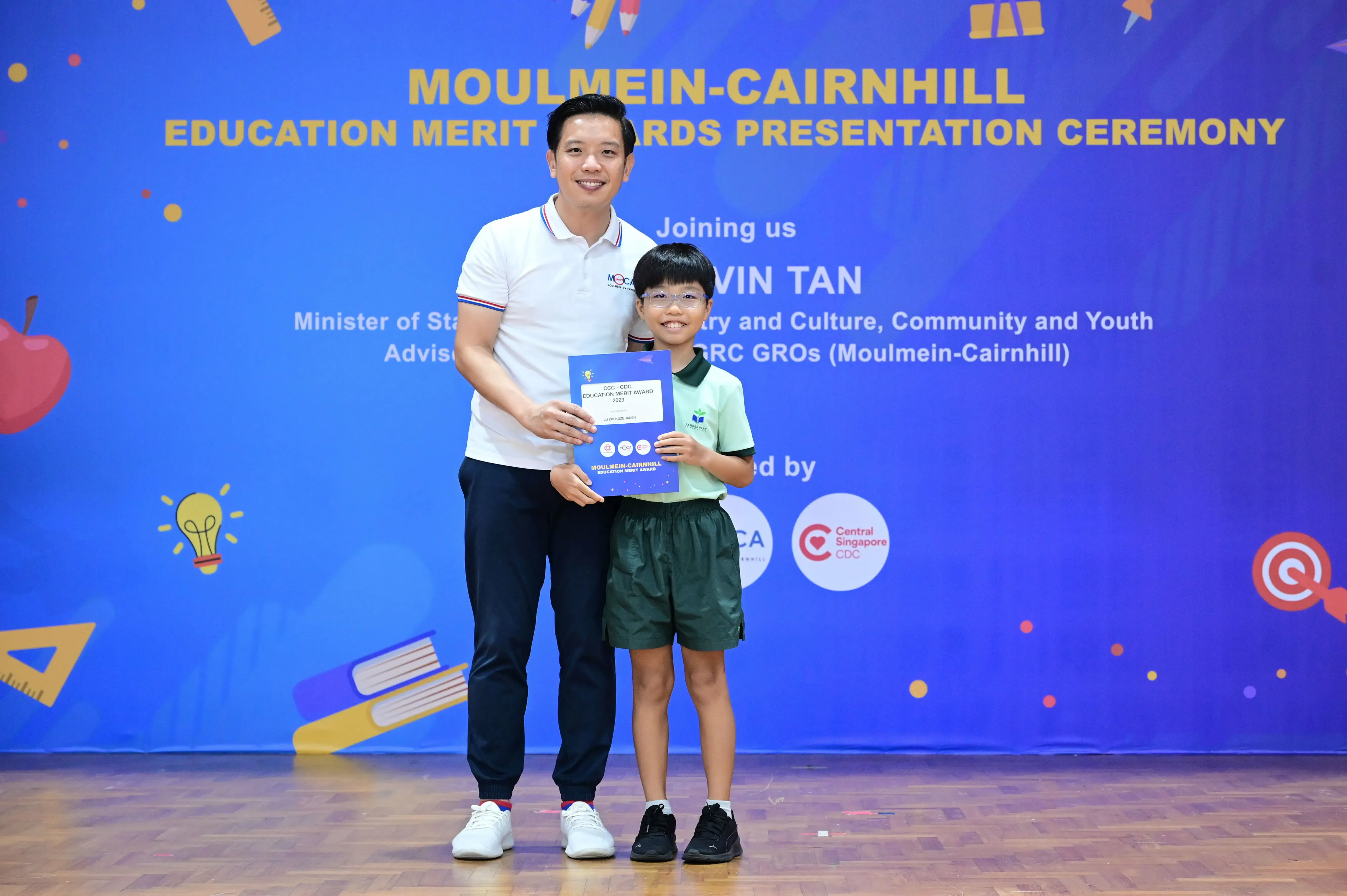 An adult and a young boy hold a certificate in front of a Moulmein-Cairnhill Education Merit Awards banner.