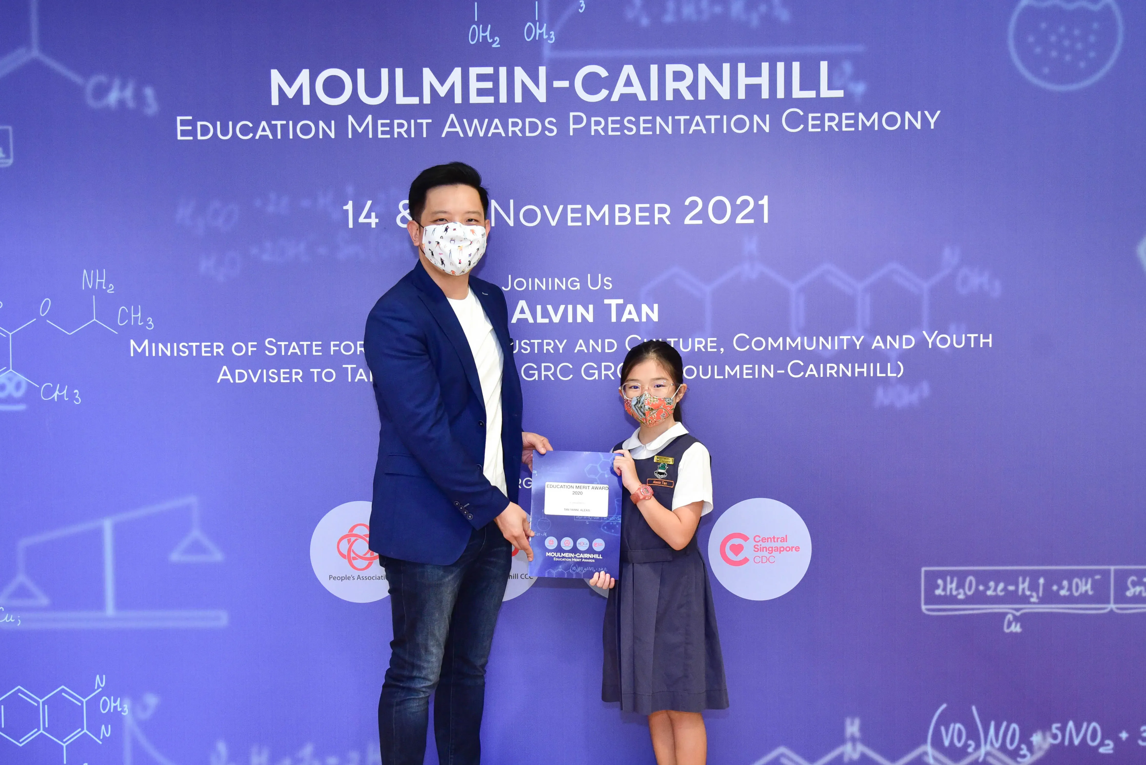 A masked man and a young girl in school uniform hold an award certificate at the Moulmein-Cairnhill Education Merit Awards 2021.