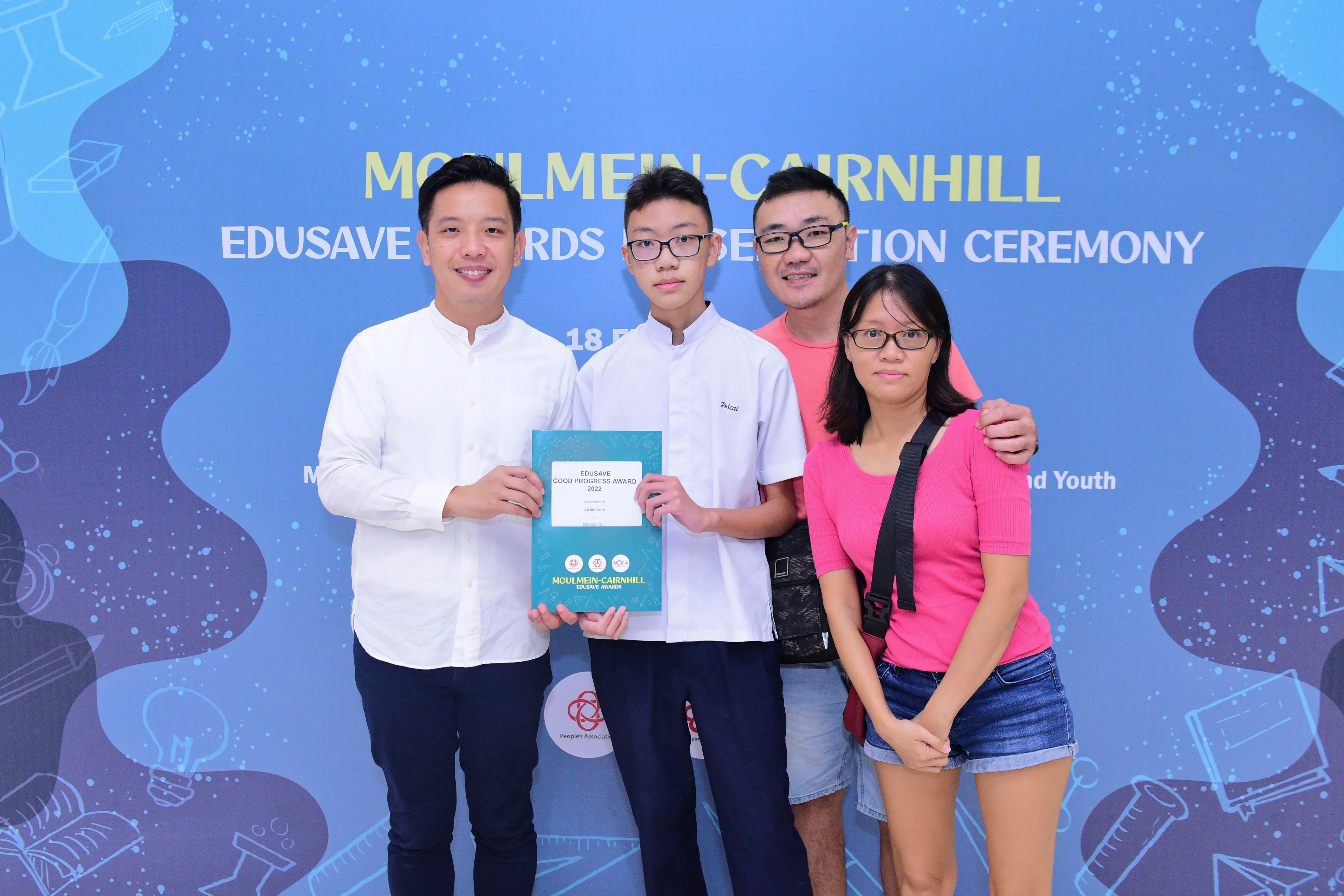 Four people pose smiling before a Moulmein-Cairnhill Edusave Awards banner, with a boy holding a teal award certificate.