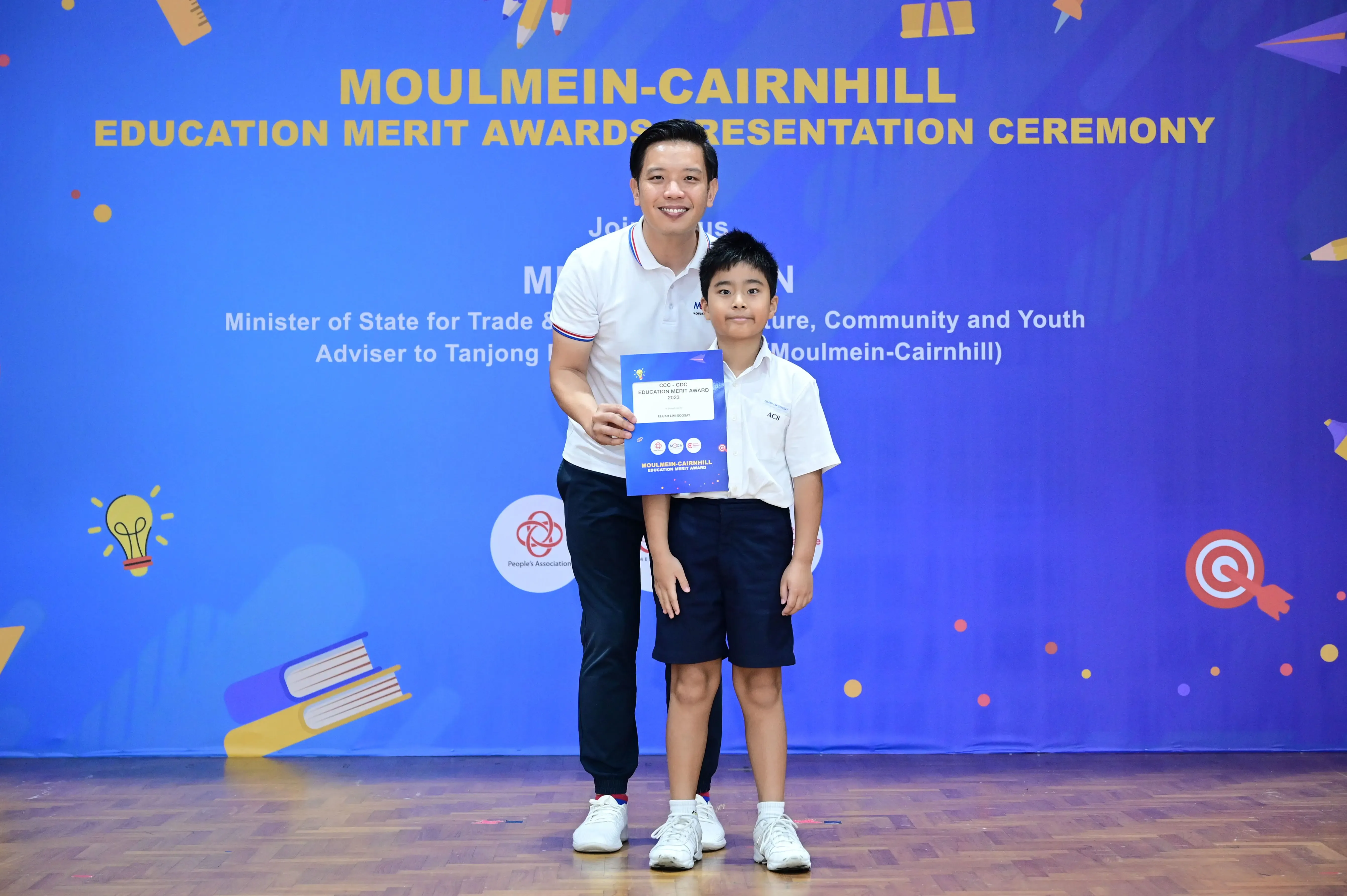 An adult and a young boy hold an award certificate in front of a Moulmein-Cairnhill Education Merit Awards banner.