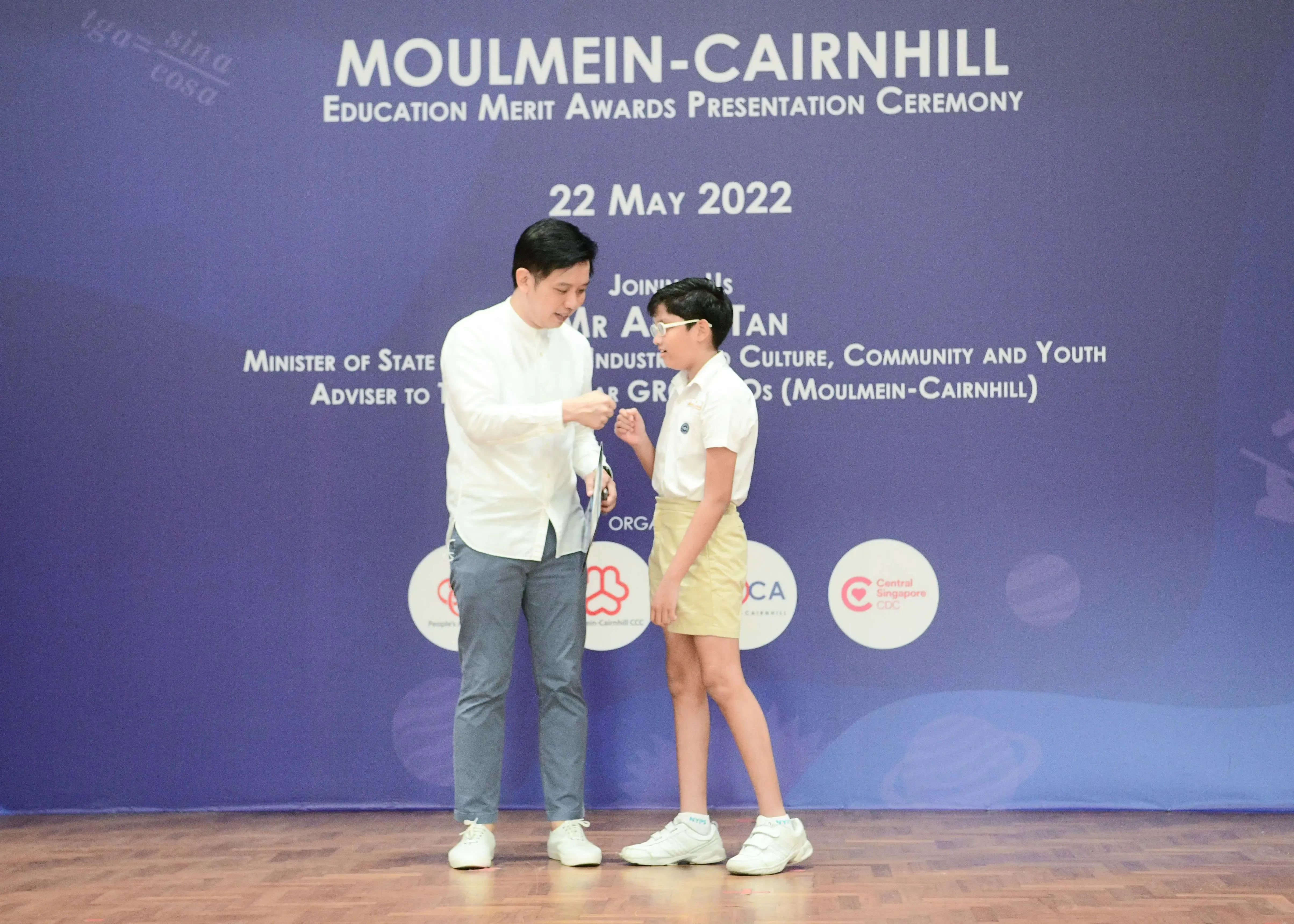 A man in white presents an award to a young student on stage at the Moulmein-Cairnhill Education Merit Awards, 22 May 2022.