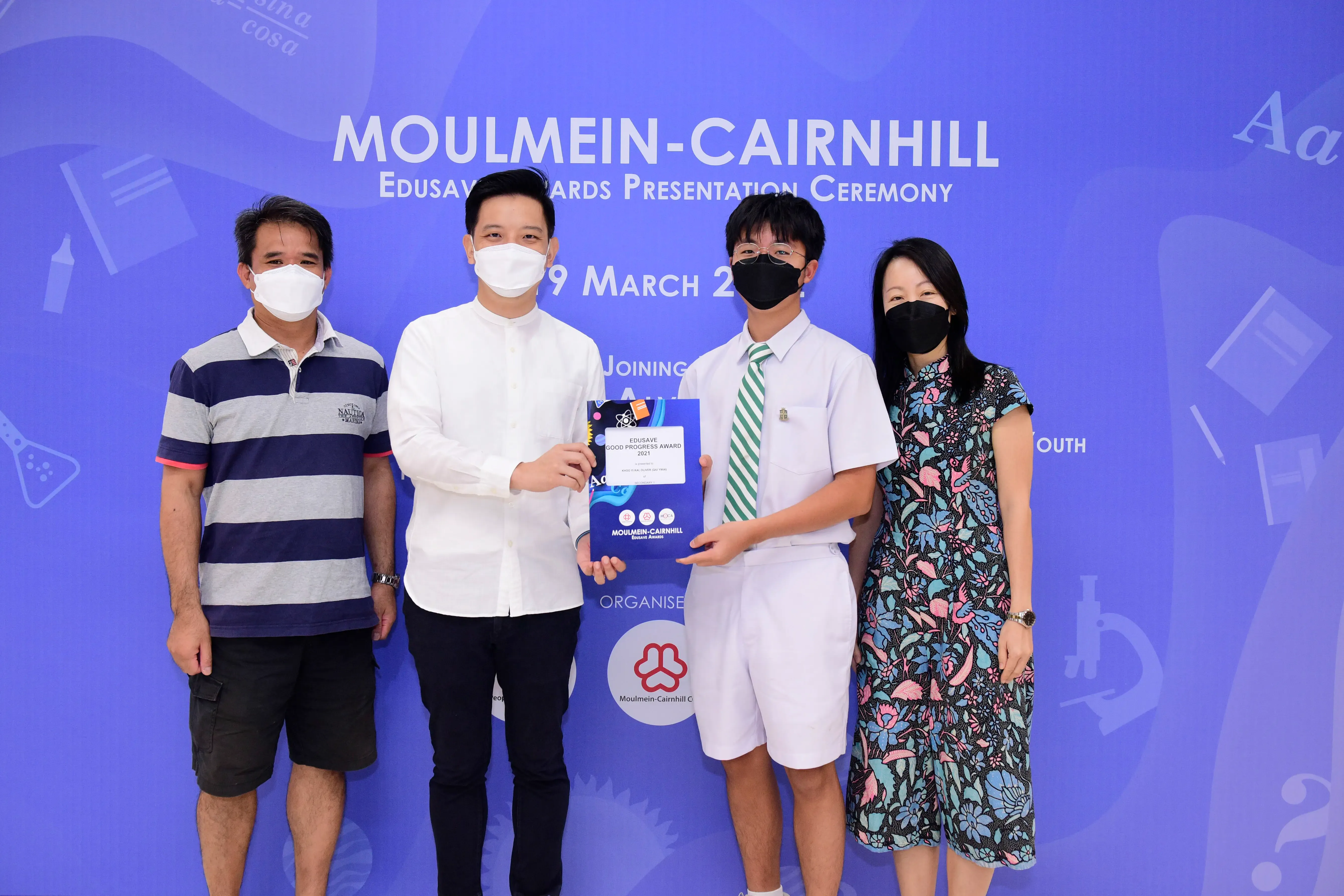 Four masked people pose at the Moulmein-Cairnhill Edusave Awards Presentation Ceremony, with two holding a certificate.