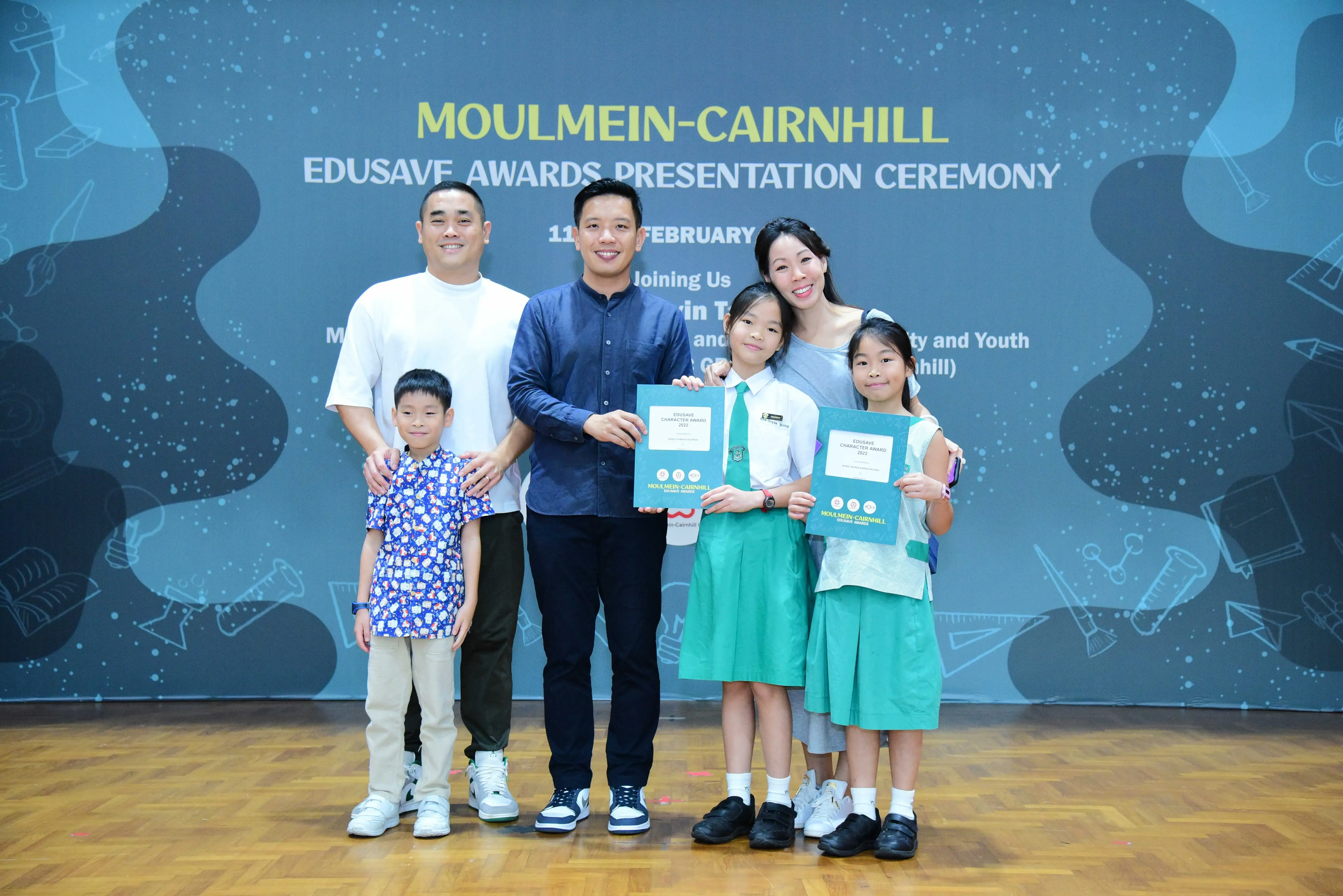 Six people pose in front of a Moulmein-Cairnhill Edusave Awards Presentation Ceremony banner, two girls holding certificates.