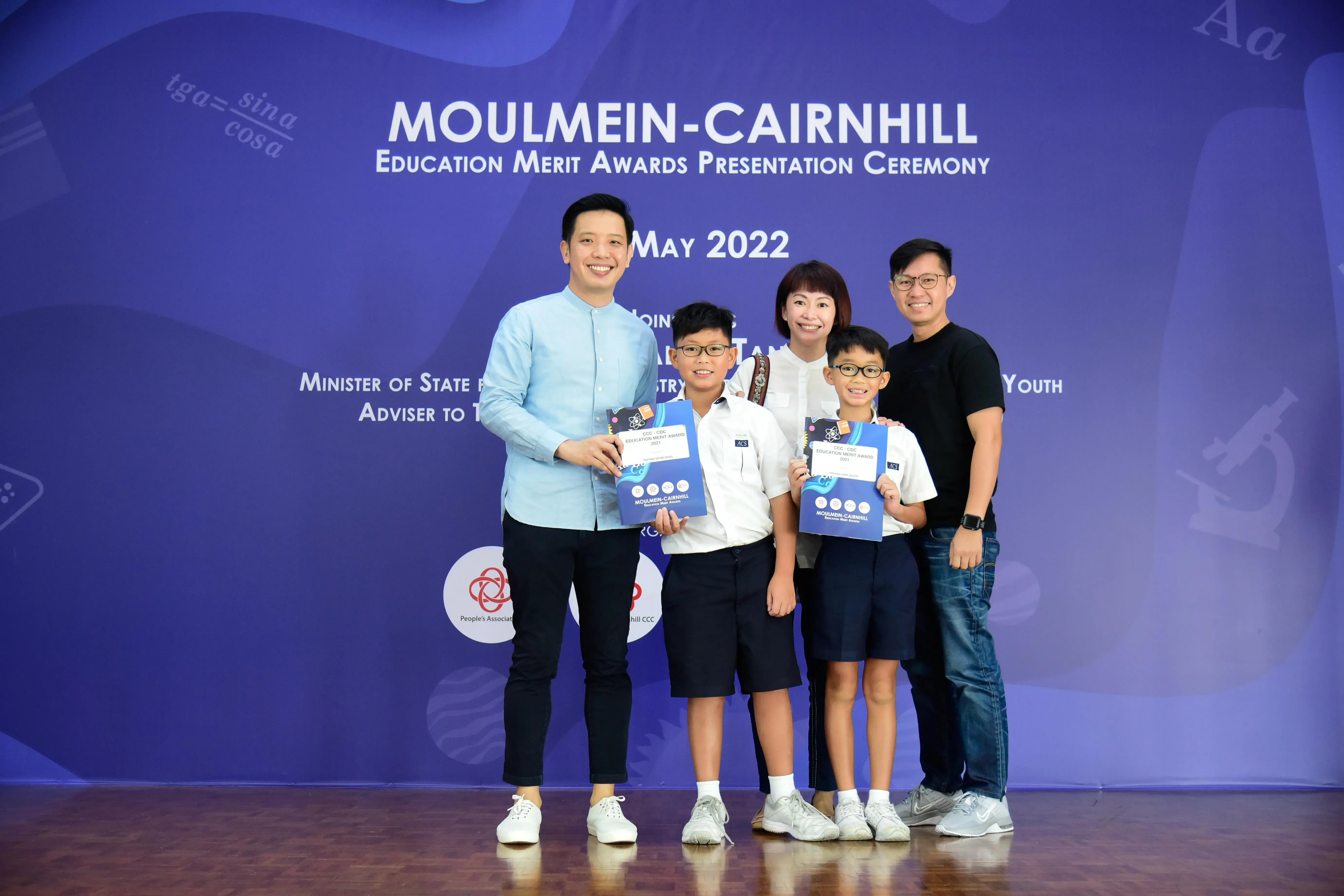 Five people pose at the Moulmein-Cairnhill Education Merit Awards Ceremony May 2022, two boys holding award booklets.