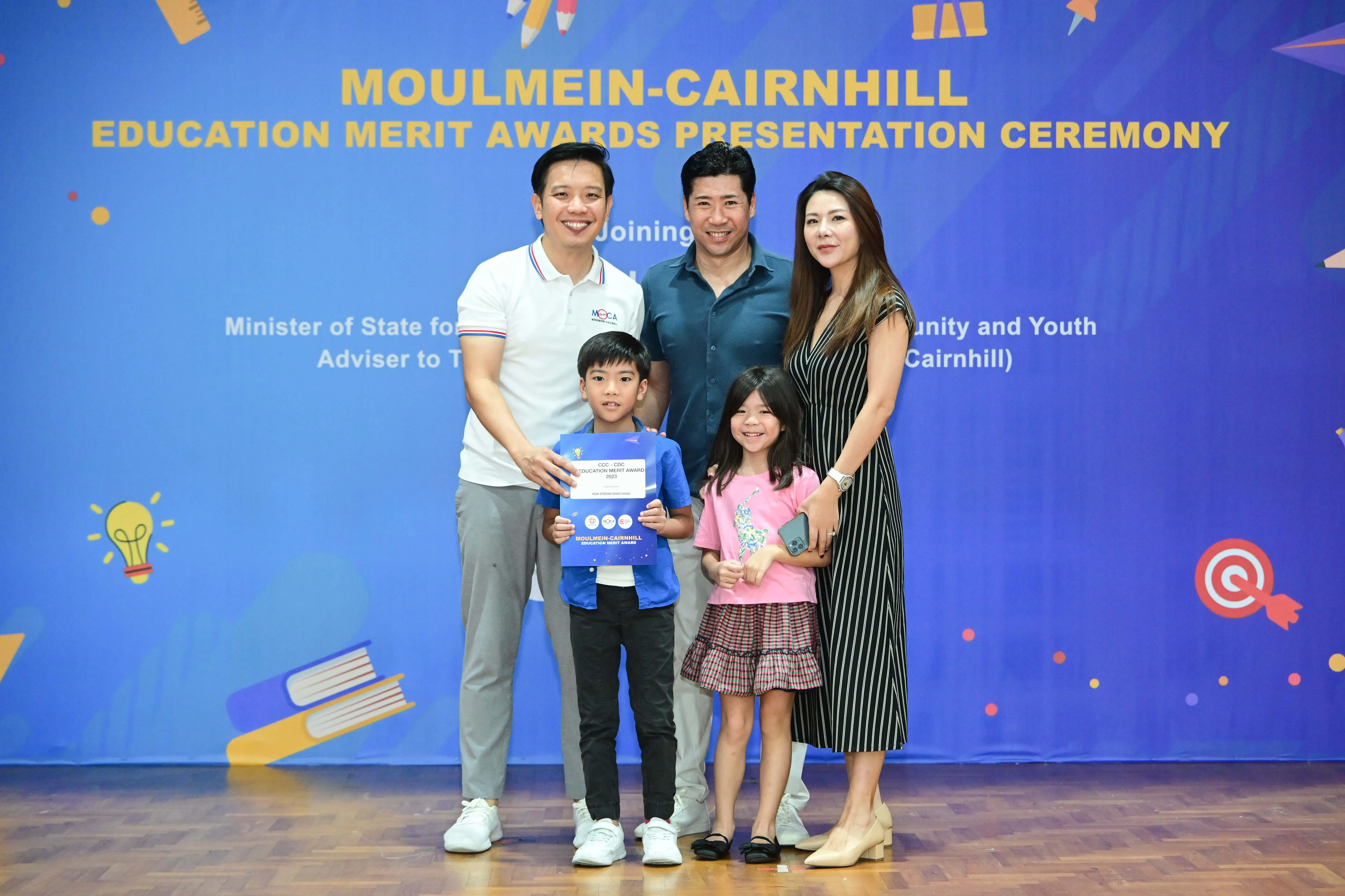 Three adults and two children pose at the Moulmein-Cairnhill Education Merit Awards Presentation Ceremony, boy holds certificate.