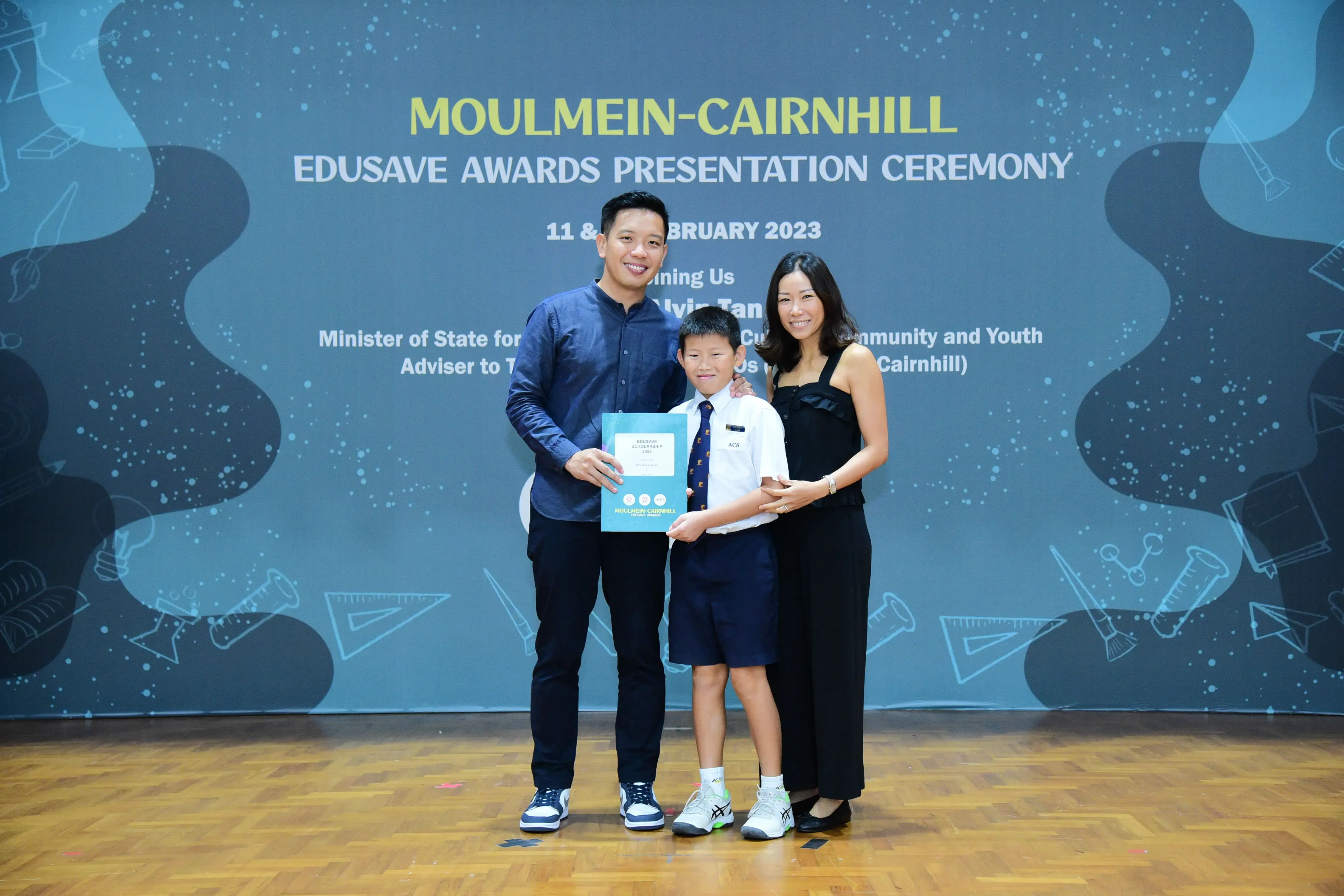 A boy in school uniform holds a certificate flanked by two adults at the Moulmein-Cairnhill Edusave Awards 2023.
