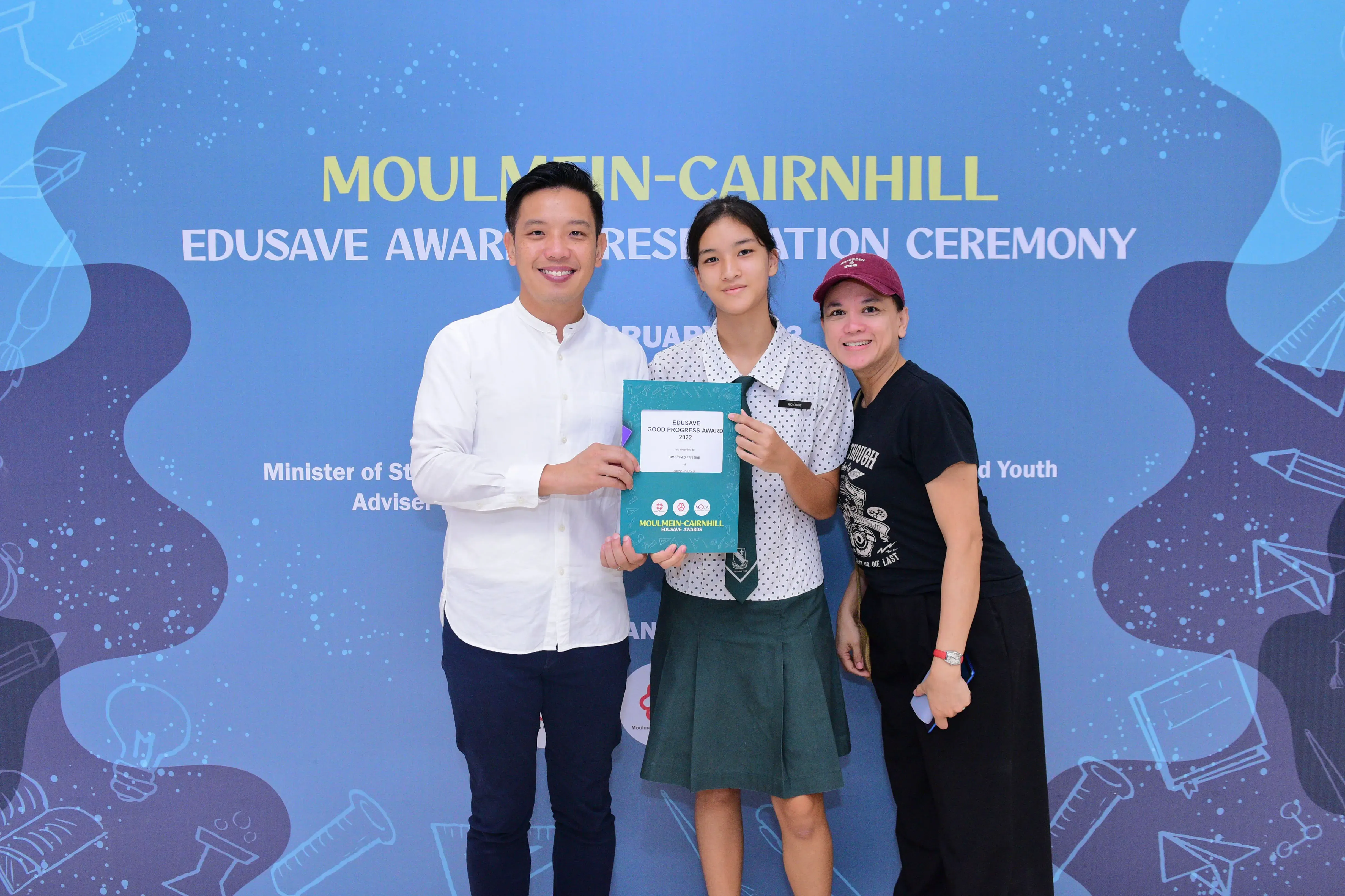 Three people pose smiling at the Moulmein-Cairnhill Edusave Awards Presentation Ceremony, with a student holding an award.