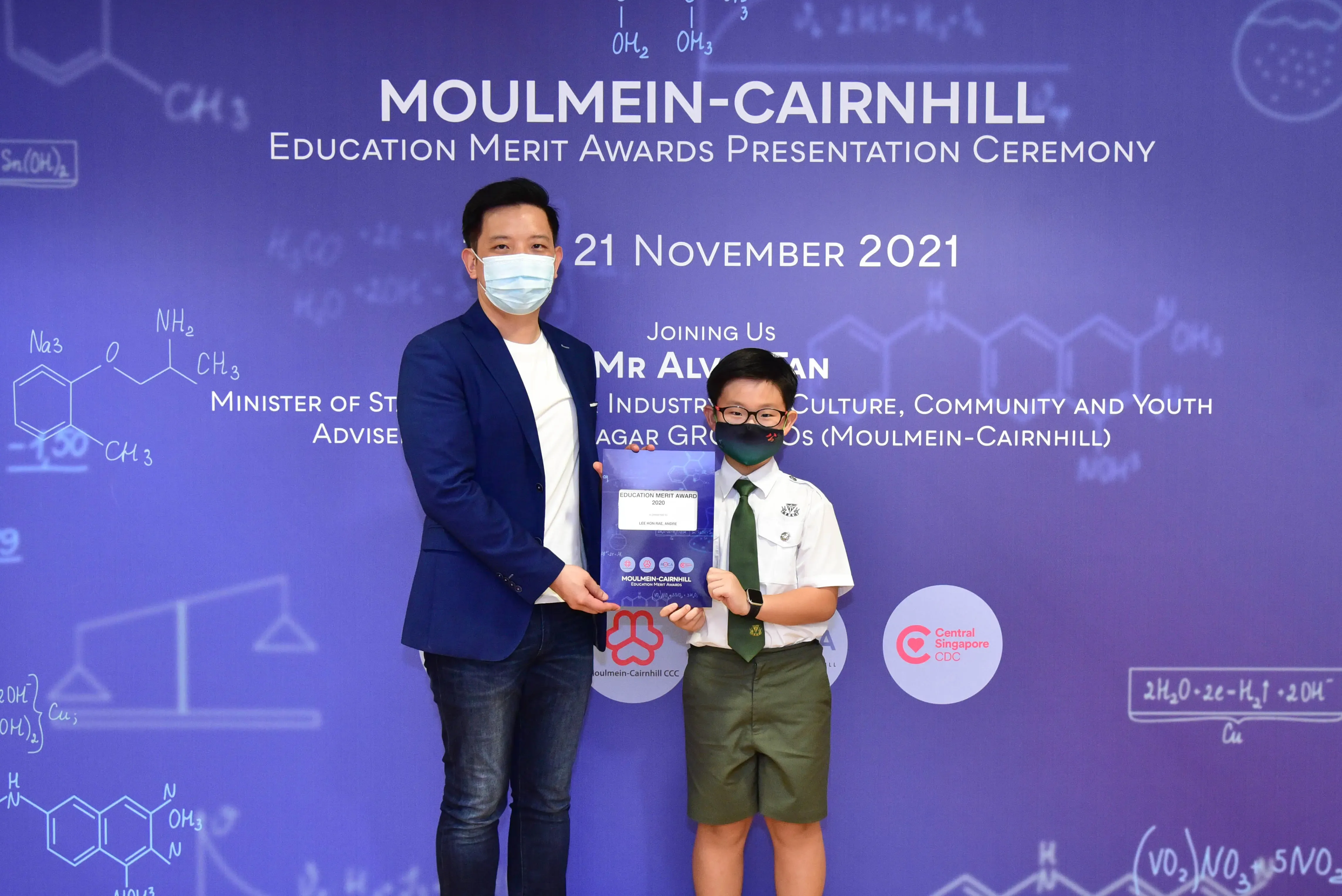 An adult and a young student in uniform hold an award at the Moulmein-Cairnhill Education Merit Awards ceremony, November 2021.