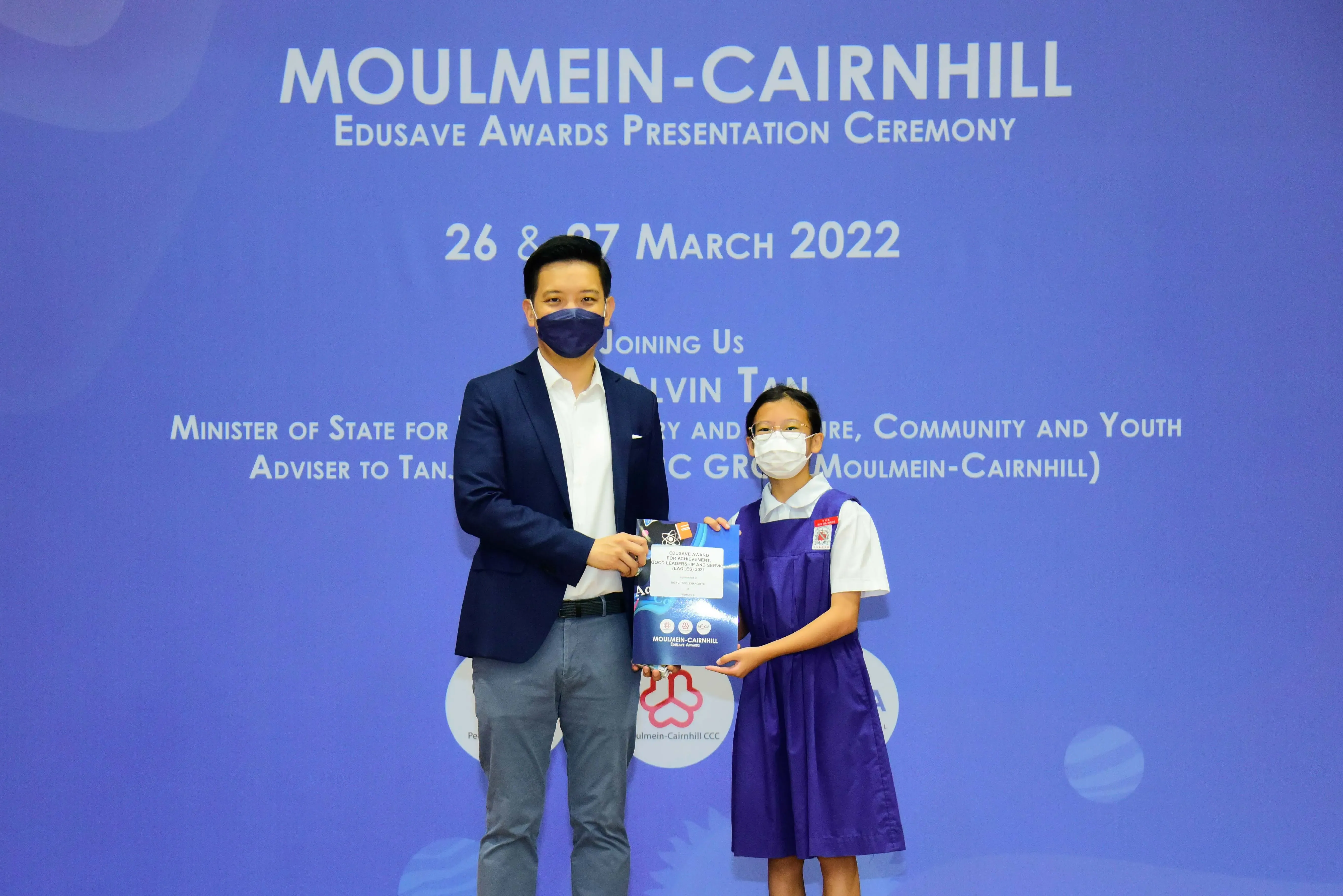 A masked man in a suit and a young girl in school uniform hold an award at the Moulmein-Cairnhill Edusave ceremony.