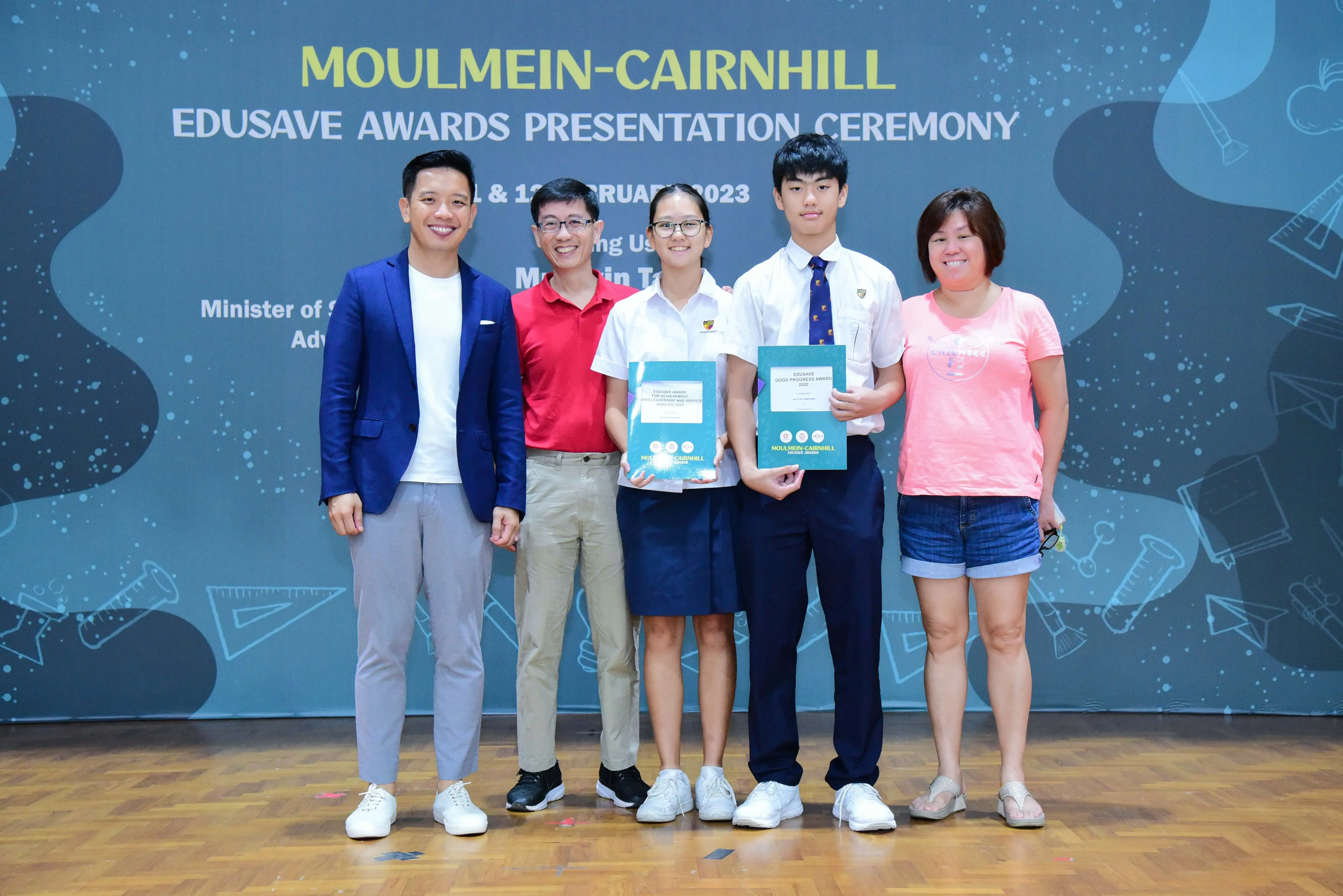 Five people pose at the Moulmein-Cairnhill Edusave Awards Presentation Ceremony 2023, two students holding teal award booklets.
