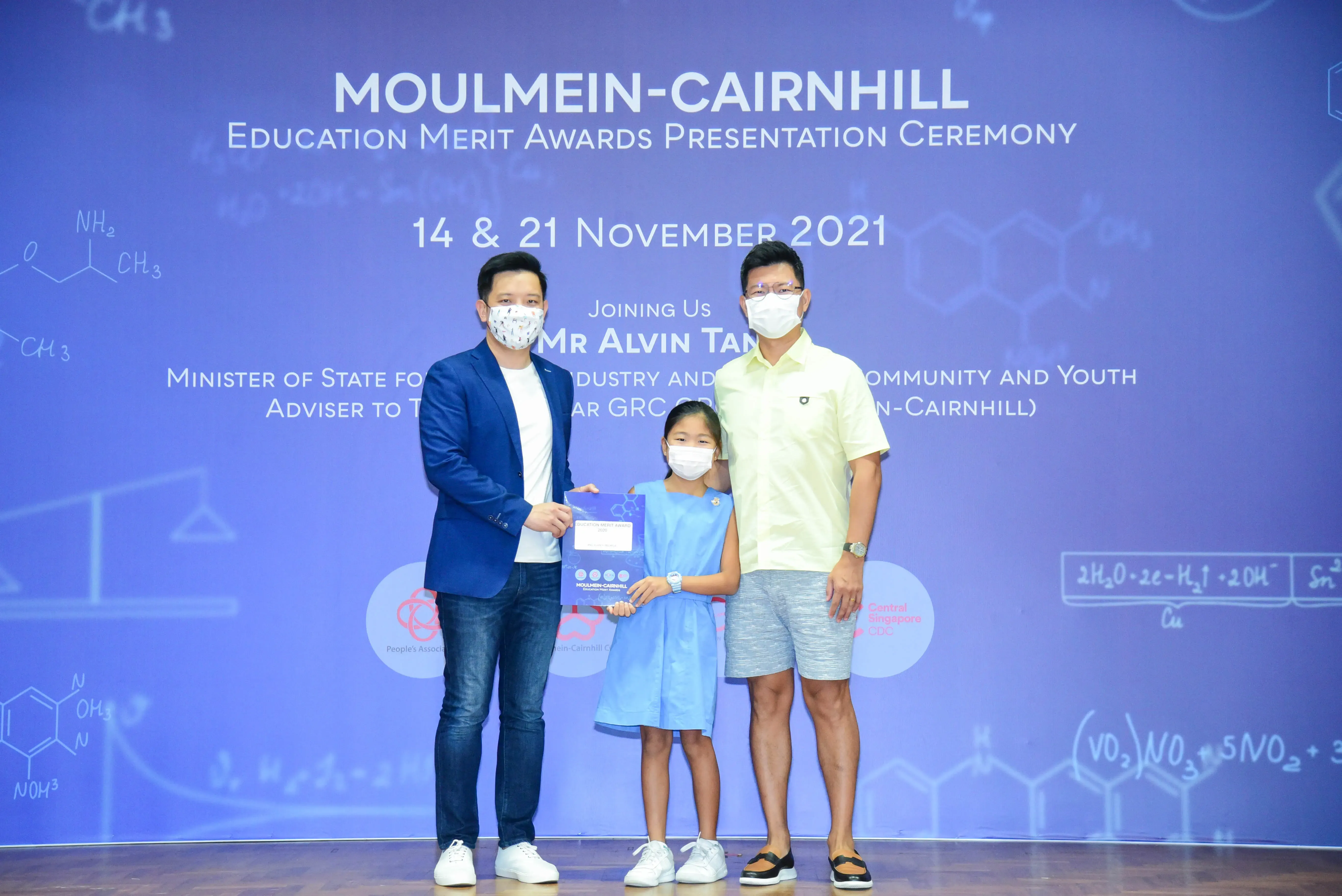 Two masked men and a young girl holding an award at the Moulmein-Cairnhill Education Merit Awards Ceremony 2021.