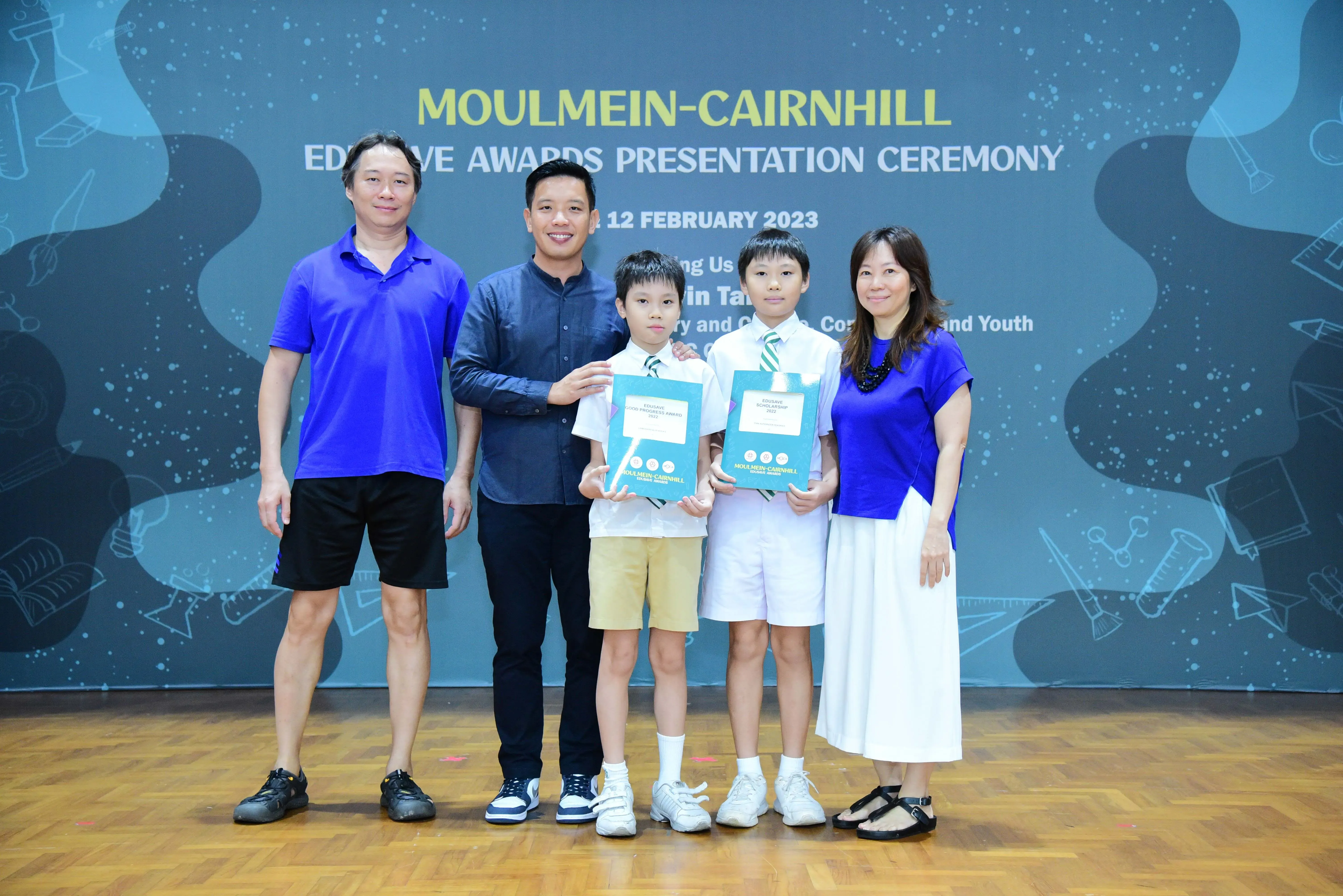Two adults and two uniformed children hold award folders at the Moulmein-Cairnhill Education Awards Ceremony, February 2023.